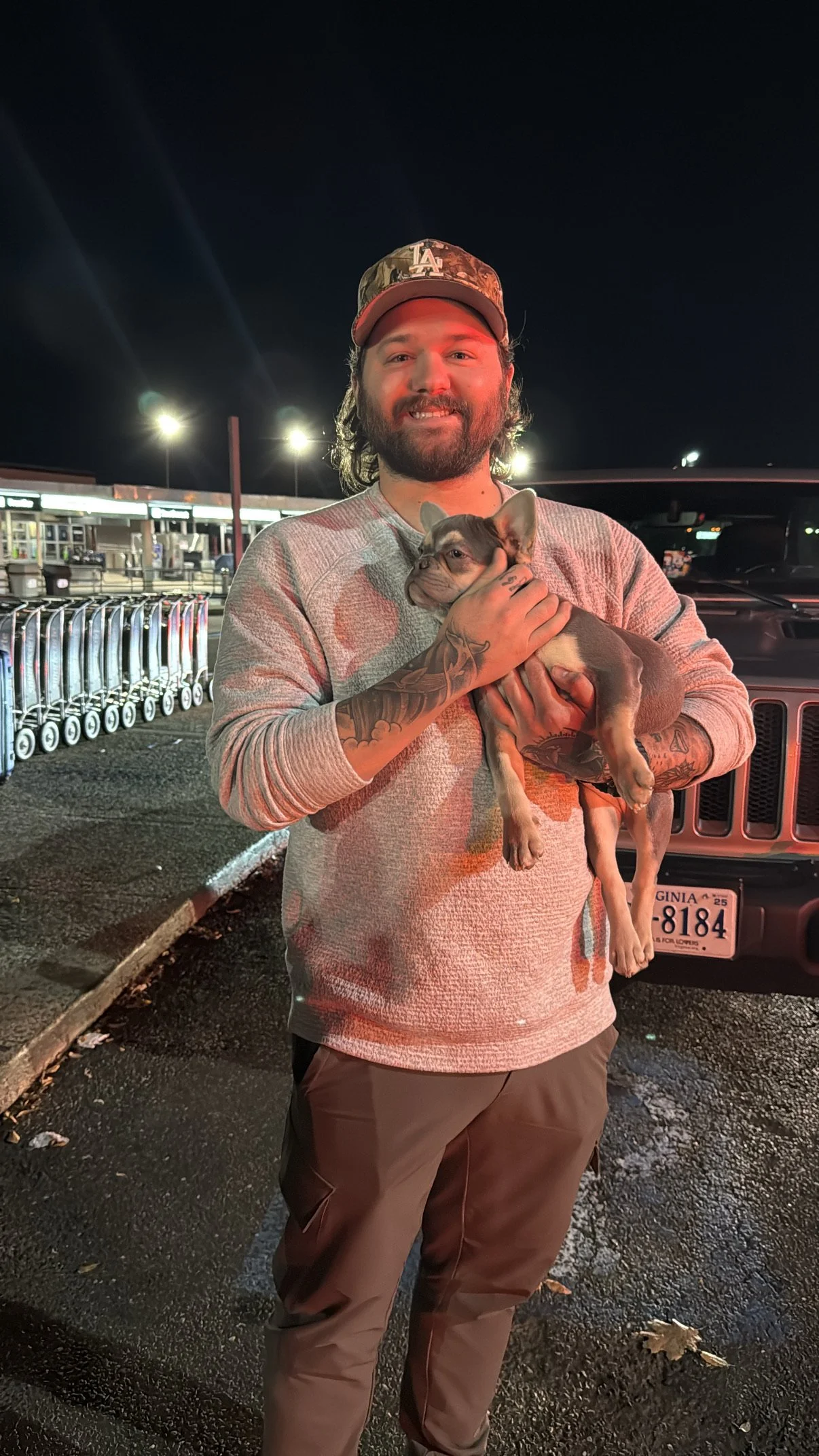 A man with tattoos holding a French Bulldog puppy at night in a parking lot with shopping carts and headlights in the background.