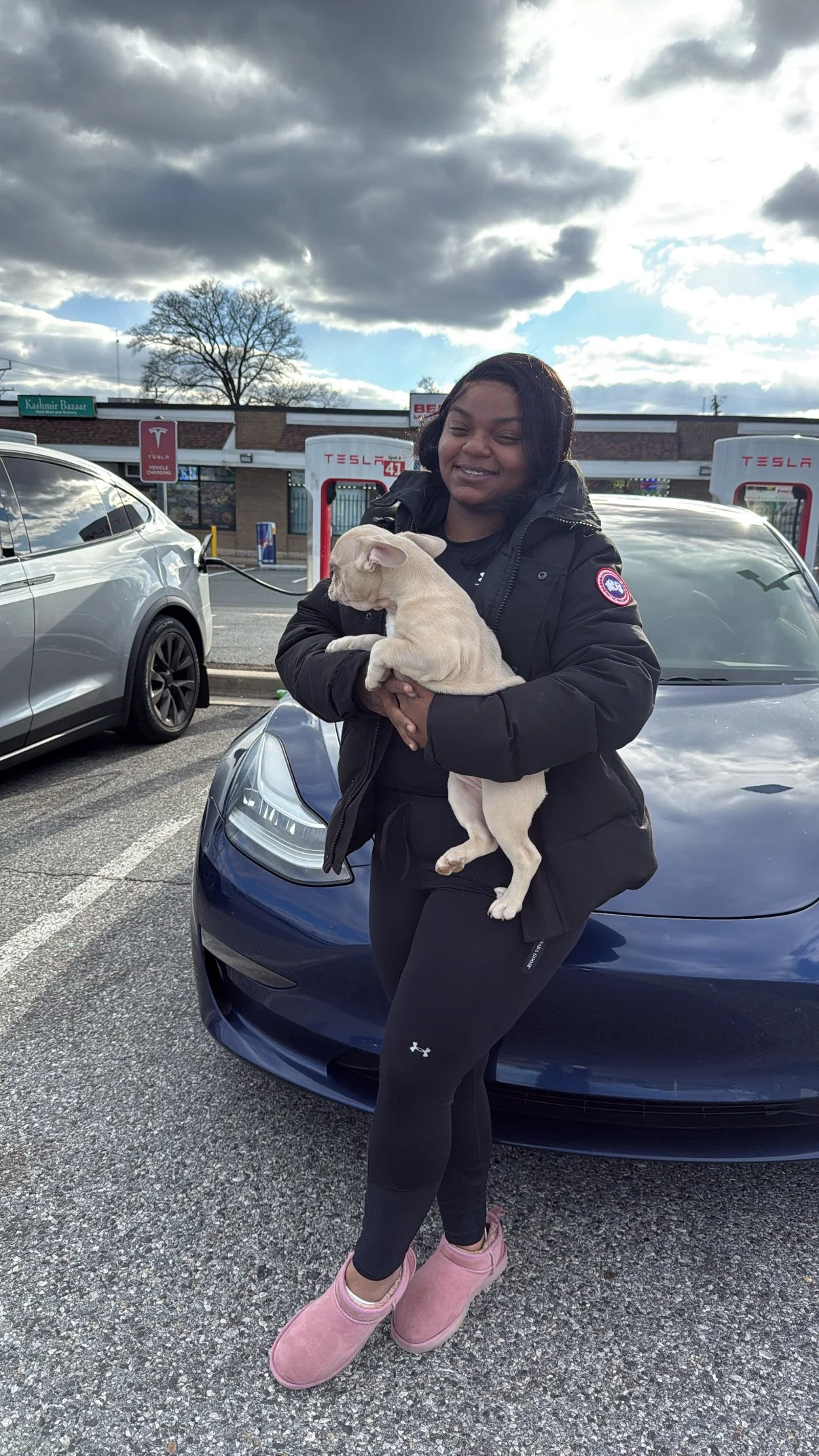 A woman standing in a parking lot holding a small tan dog, smiling with a cloudy sky above and Teslas charging at station behind her.