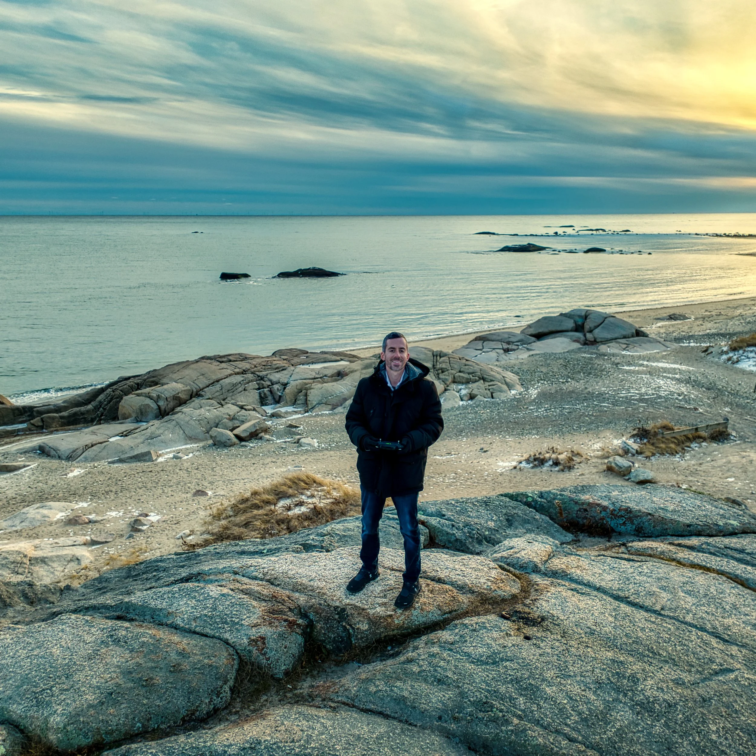 A man standing on rocks near the beach during sunset or sunrise with a calm ocean and cloudy sky in the background.