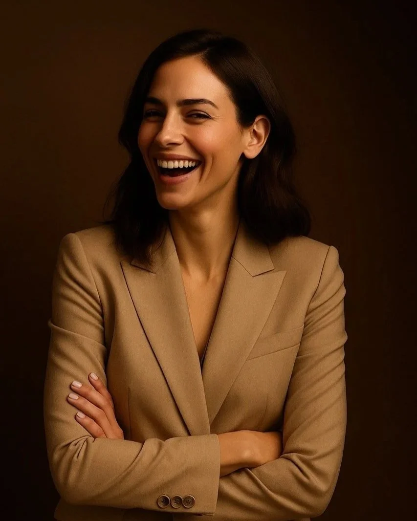 A woman with dark hair wearing a tan blazer, smiling, and crossing her arms against a dark brown background.