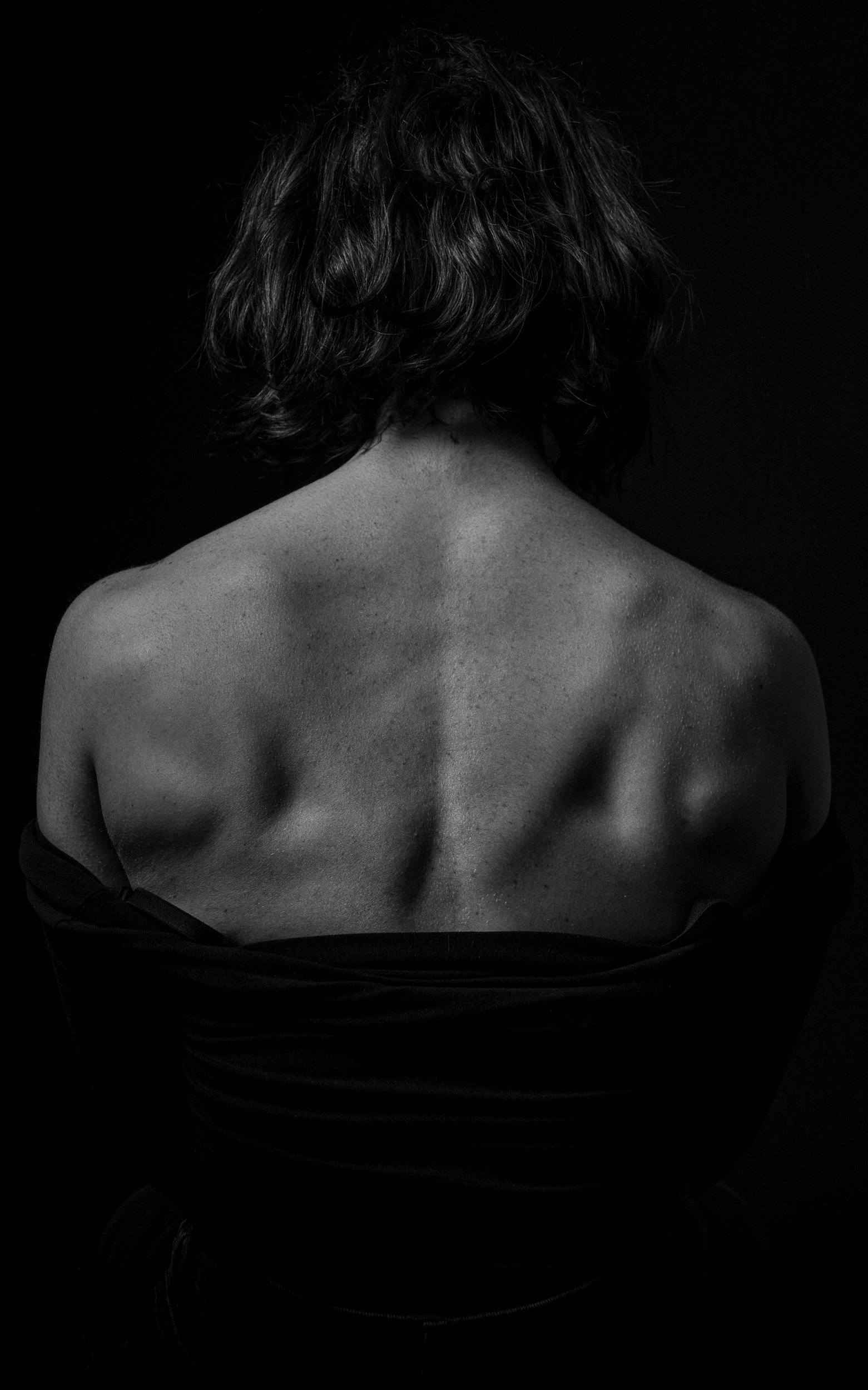Back view of a woman with curly short hair, topless, with her arms crossed in front of her, against a black background.