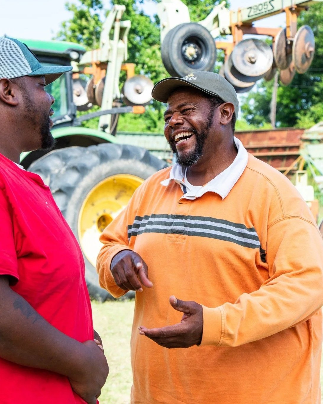 Two Black men having a conversation outdoors, one wearing a red t-shirt and gray cap, the other in an orange striped shirt and gray cap, smiling and gesturing, with farming equipment and a tractor in the background.