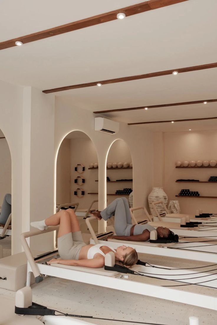 Two women lying on Pilates reformer machines in a wellness studio, engaging in a fitness or Pilates session.