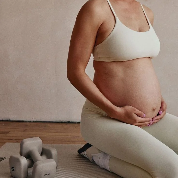 A pregnant woman in a beige sports bra and cream-colored leggings sitting on the floor in a yoga pose, holding her belly with both hands. There are two small dumbbells on the mat nearby, in a room with wooden floors and a plain wall.
