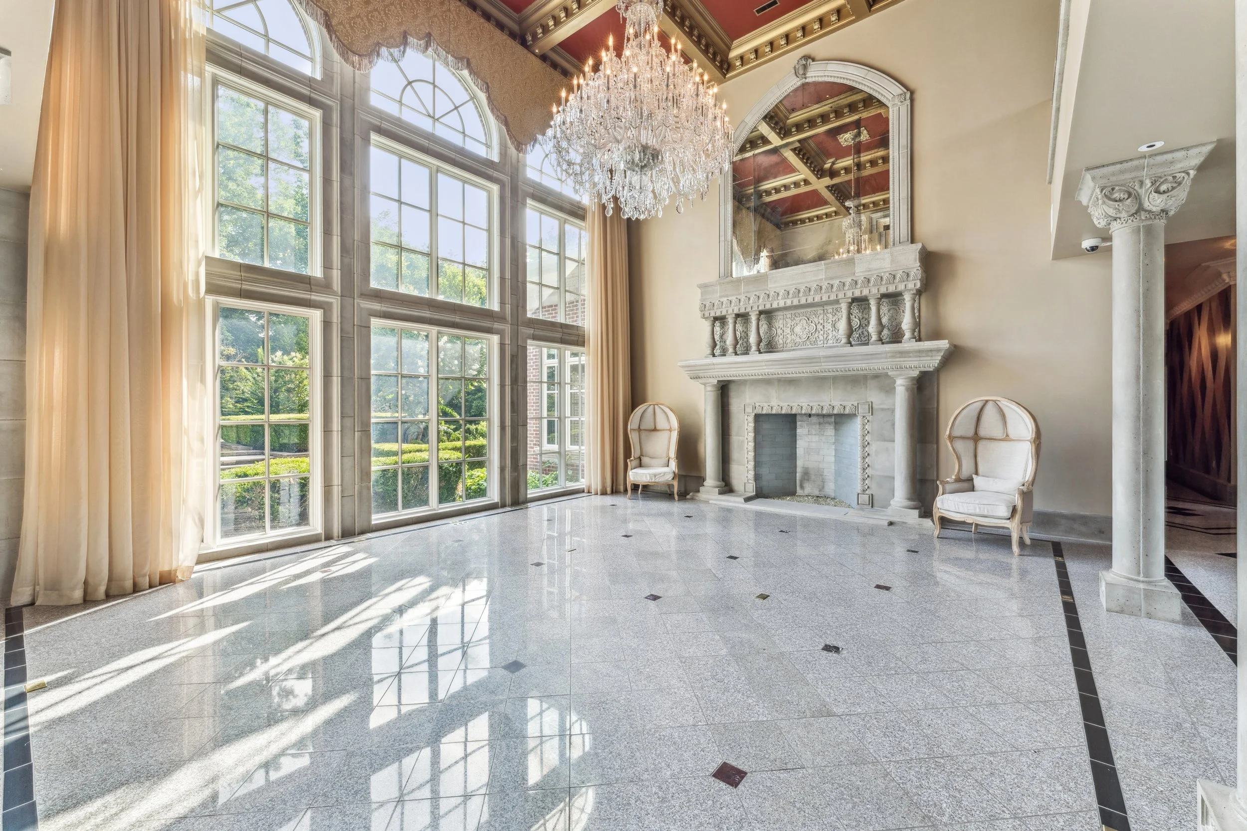 Elegant interior room with large windows, a chandelier, a marble fireplace with decorative molding, and two upholstered chairs, with sunlight streaming in.