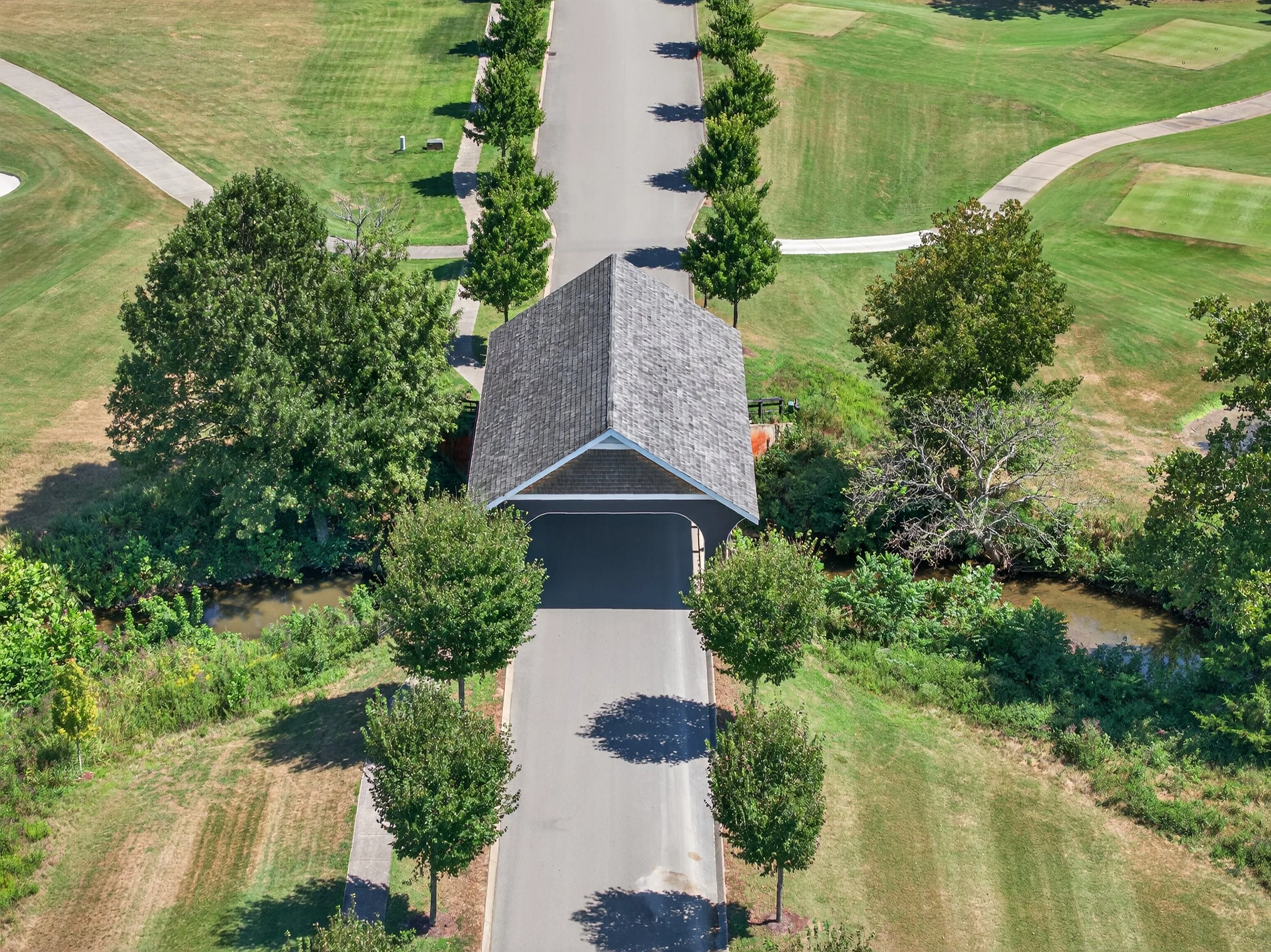 An aerial view of a park with a paved pathway lined with evenly spaced trees, surrounded by grassy areas and a building with a shingled roof.