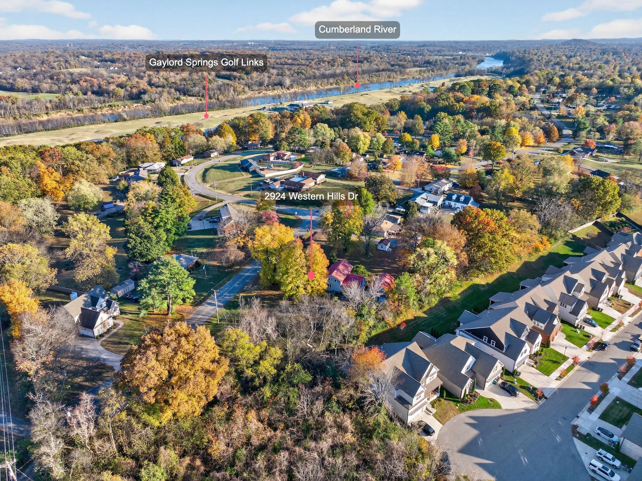 An aerial view of a suburban neighborhood with a mix of houses and trees with fall foliage, a golf course, a river labeled Cumberland River, and surrounding landscape.