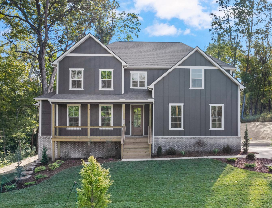 A two-story gray house with white trim and a front porch, has a brick foundation, surrounded by a green lawn, trees, and shrubs, under a blue sky with few clouds.
