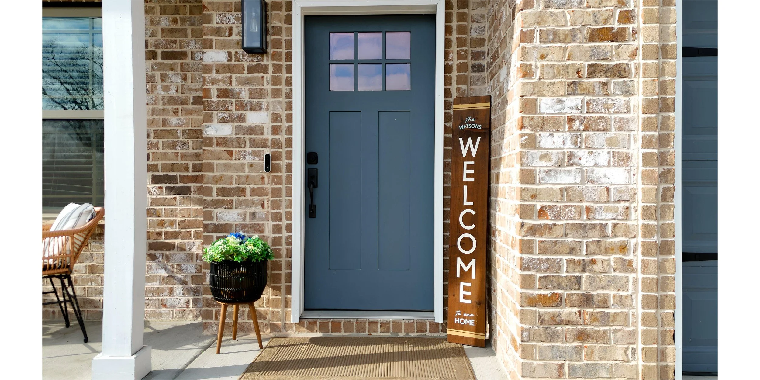 Front porch of a house with a blue door, brick walls, a wooden welcome sign, a small potted plant, and a wicker chair.