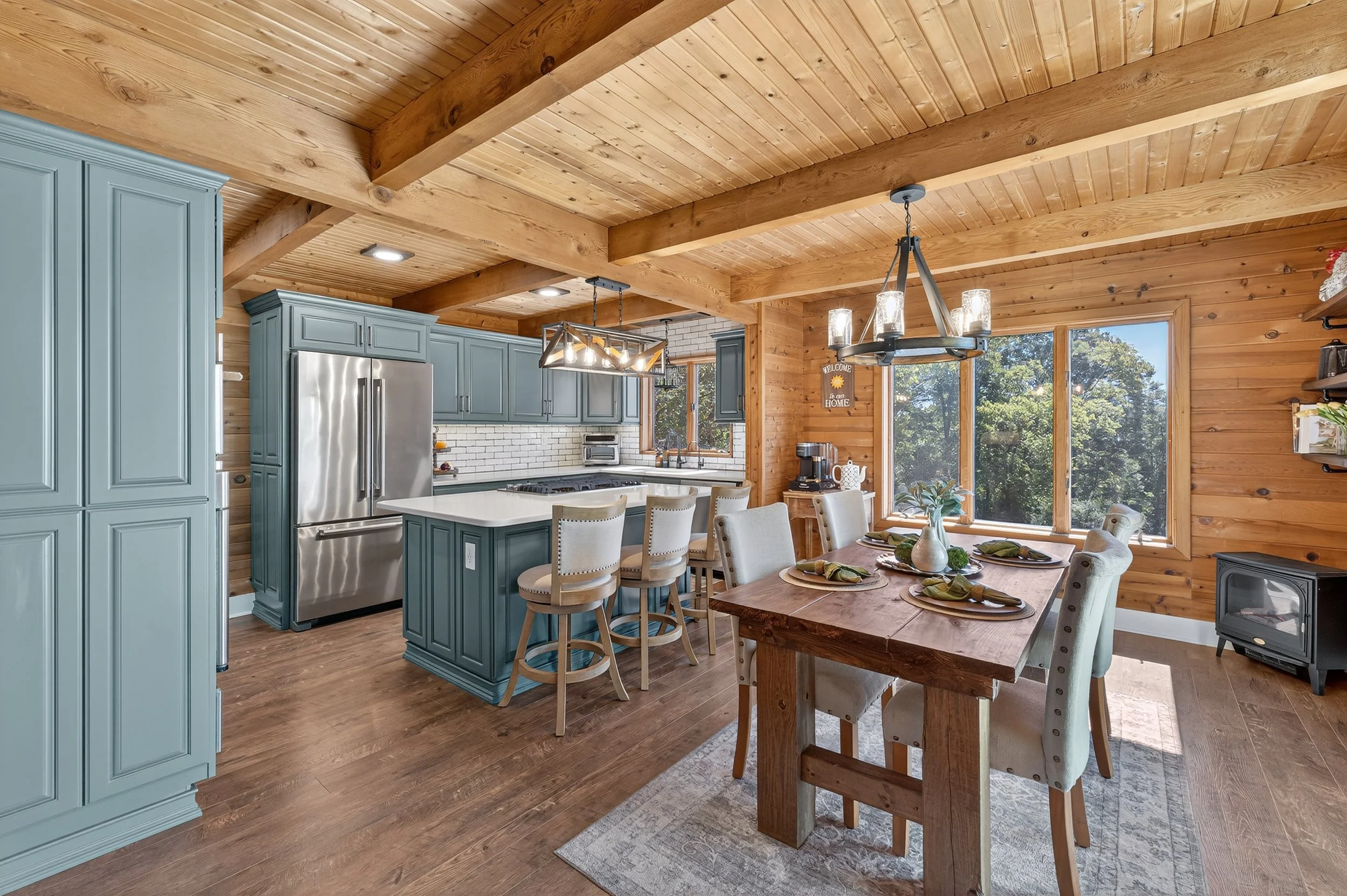 Kitchen and dining area with wooden ceiling beams and walls, blue kitchen cabinets, white countertop, stainless steel appliances, dining table with beige chairs, and large windows showing trees outside.