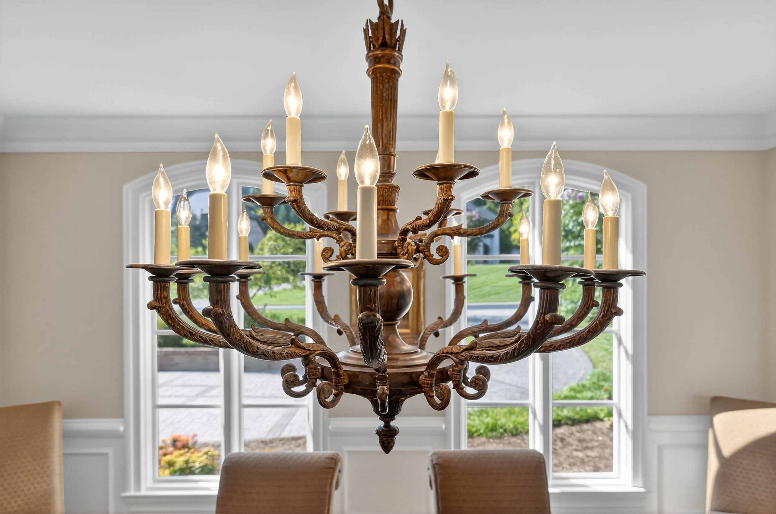 A large wooden chandelier with multiple candle-shaped lights hangs from the ceiling inside a house. Through the windows, a green lawn and trees are visible outside.