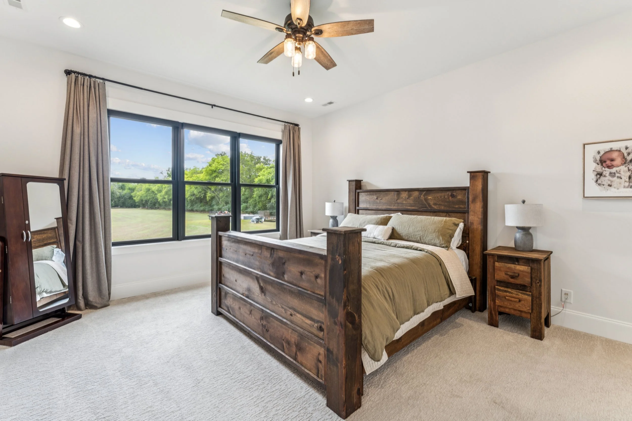 A bedroom with a wooden bed frame, beige bedding, two matching nightstands with lamps, a large window with beige curtains, a mirror, and a picture of a baby on the wall.