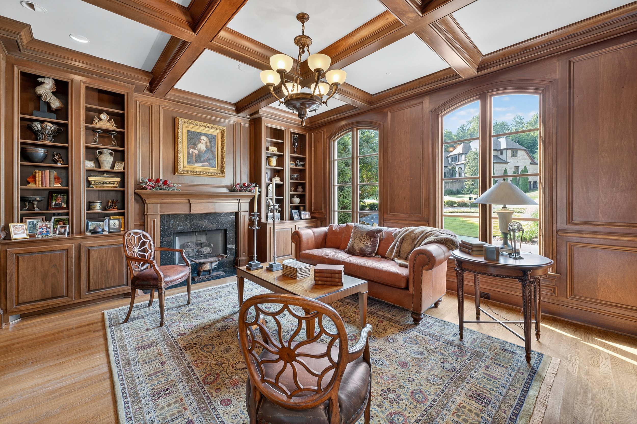 Living room with wood-paneled walls, large windows, a fireplace, and a chandelier, decorated with framed pictures, books, and a patterned rug.