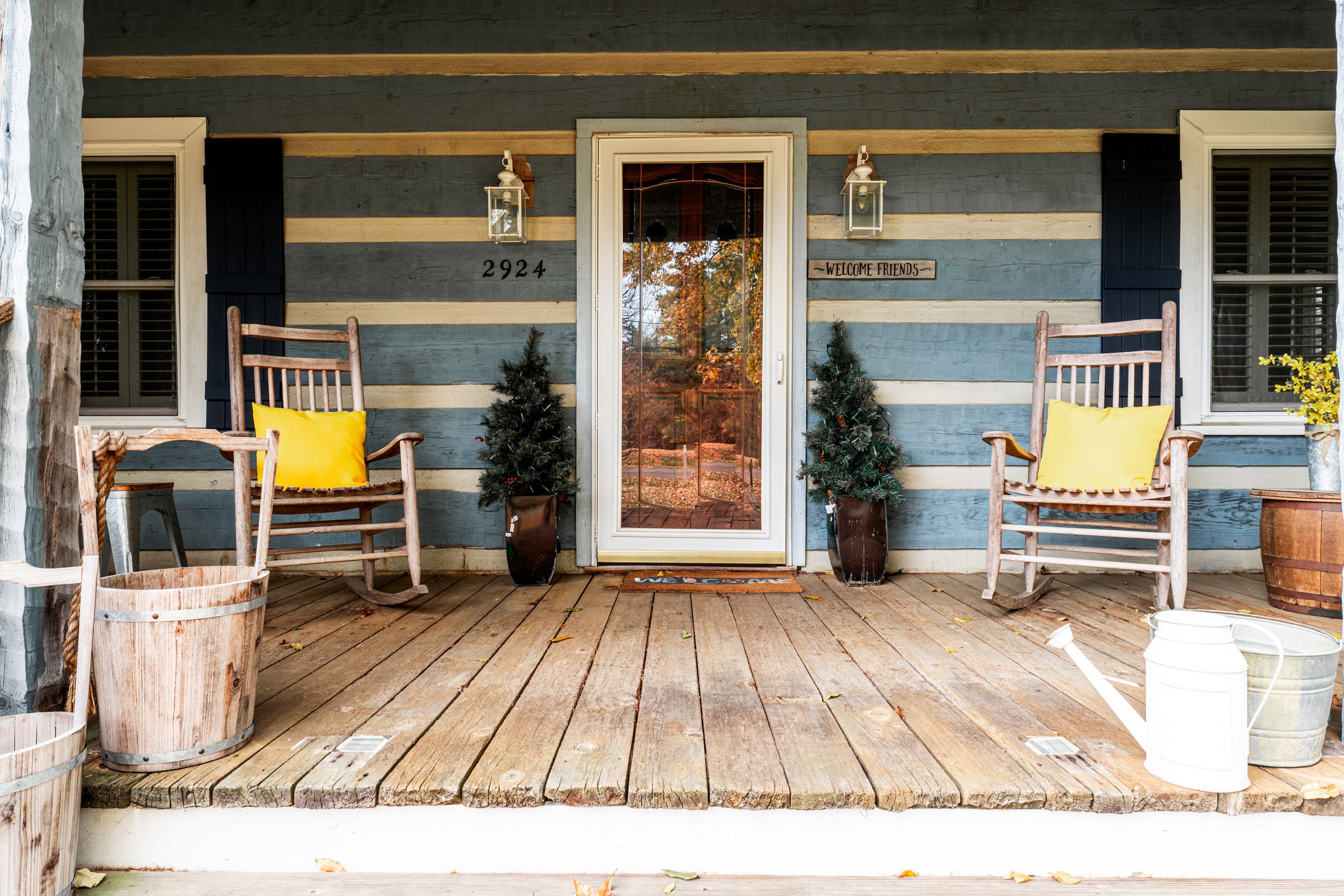 Front porch of a house with blue horizontal wooden siding, black shutters, and a glass door. Two rocking chairs with yellow pillows, small Christmas trees, wooden buckets, and a white watering can are on the porch. The house number 2924 is beside the door on the siding, and a sign on the wall reads "Welcome Friends". Reflective glass on the door shows autumn trees.