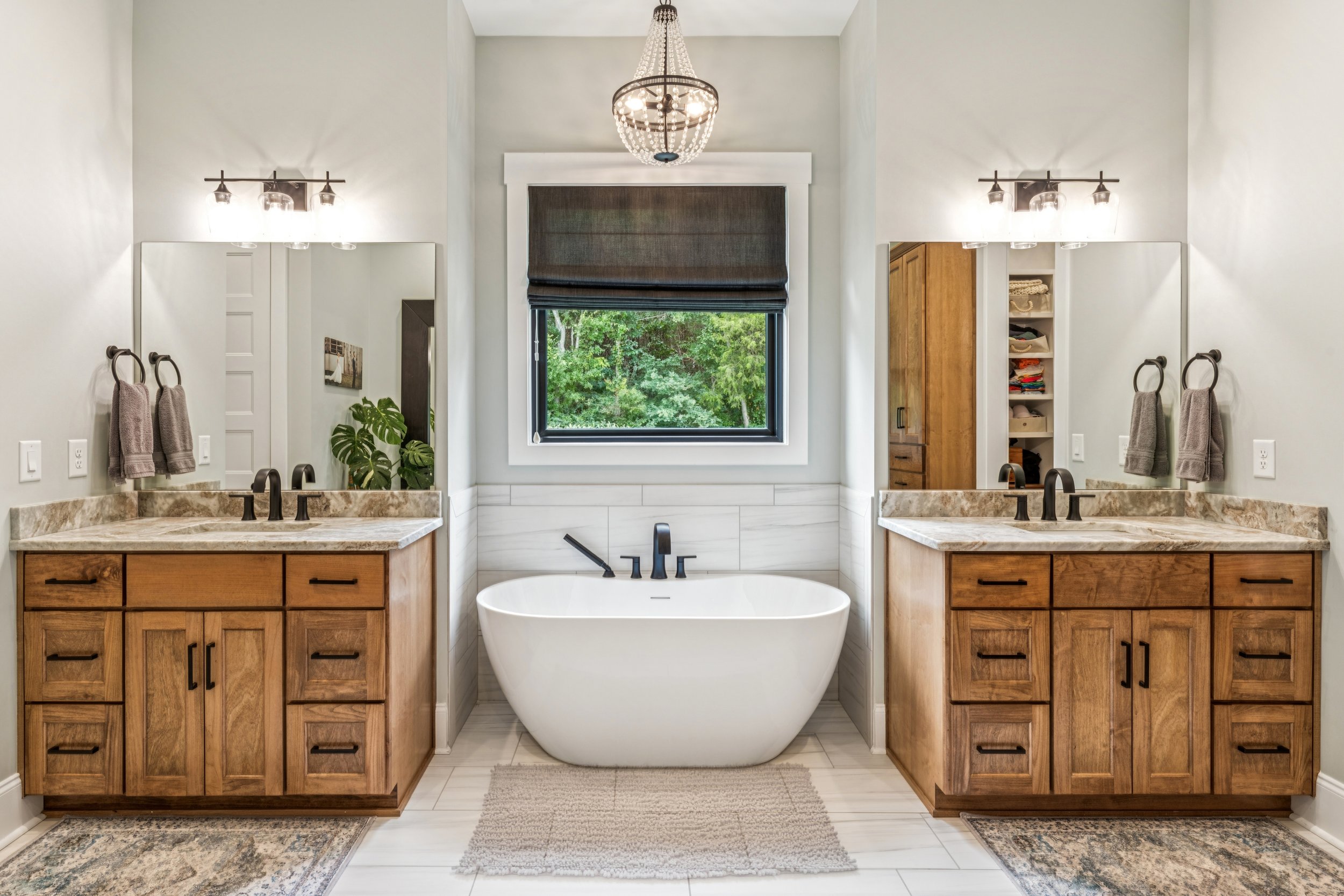 A modern bathroom featuring a central white bathtub, two wooden vanities with marble countertops, large mirrors, and a window with a dark Roman shade. There are two wall-mounted light fixtures, a chandelier hanging from the ceiling, and a plant near the mirror on the left.