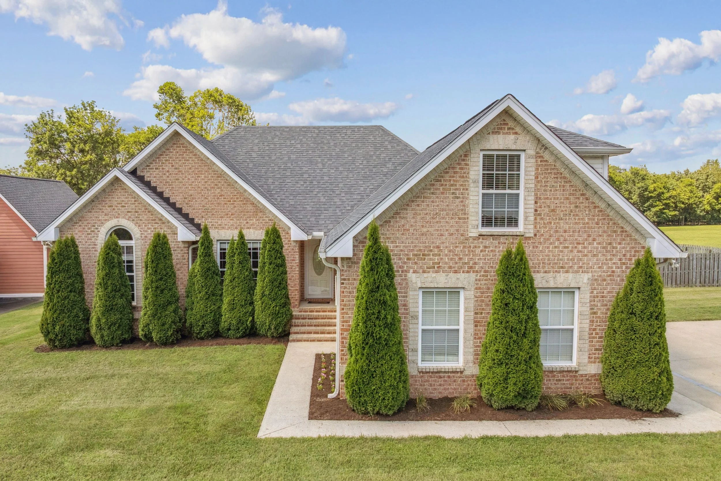 Front view of a brick house with a gray shingle roof, surrounded by green grass and tall evergreen bushes, under a blue sky with scattered white clouds.