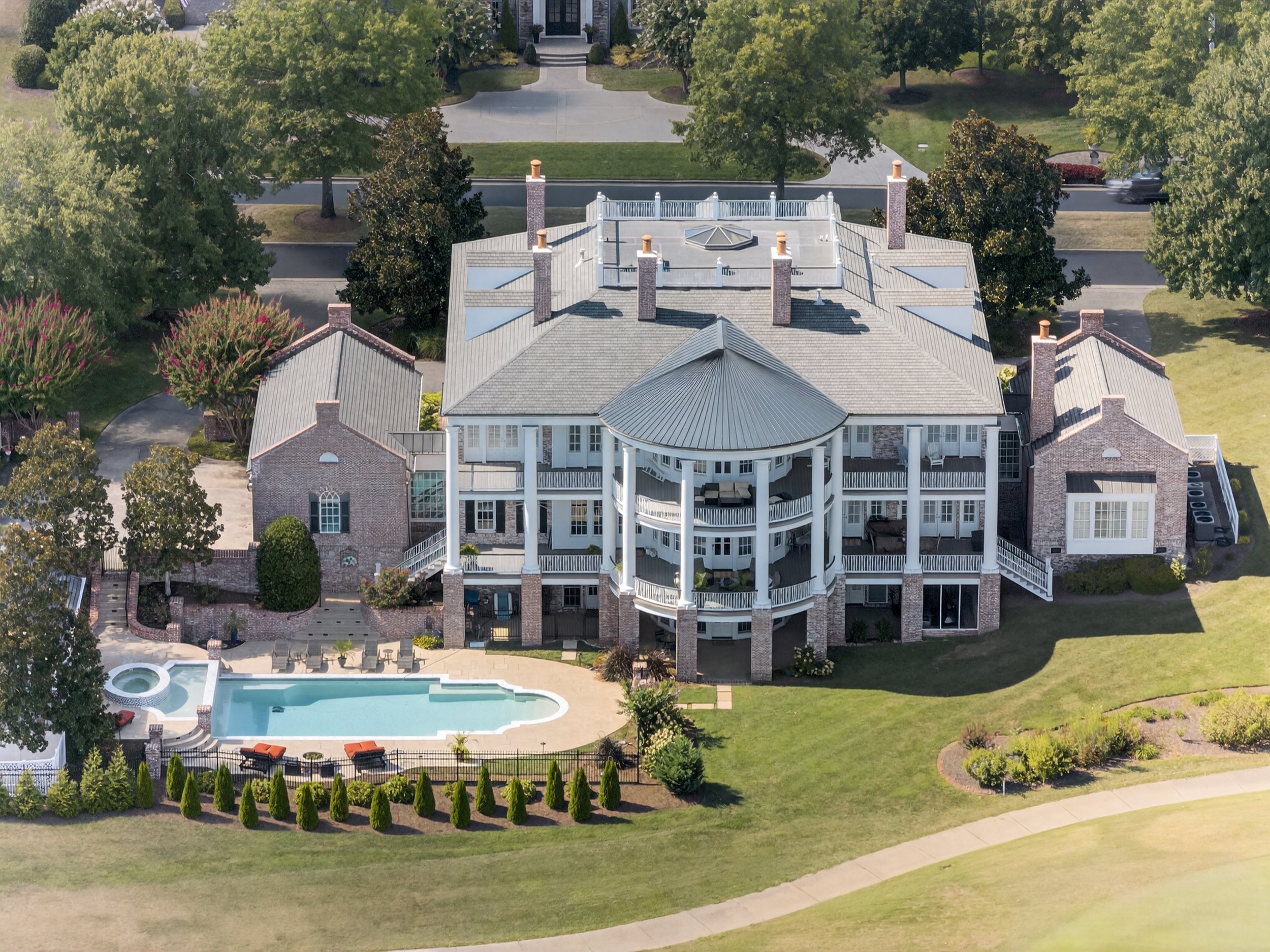 An aerial view of a large house with a pool in the backyard, surrounded by trees, a lawn, and a sidewalk.