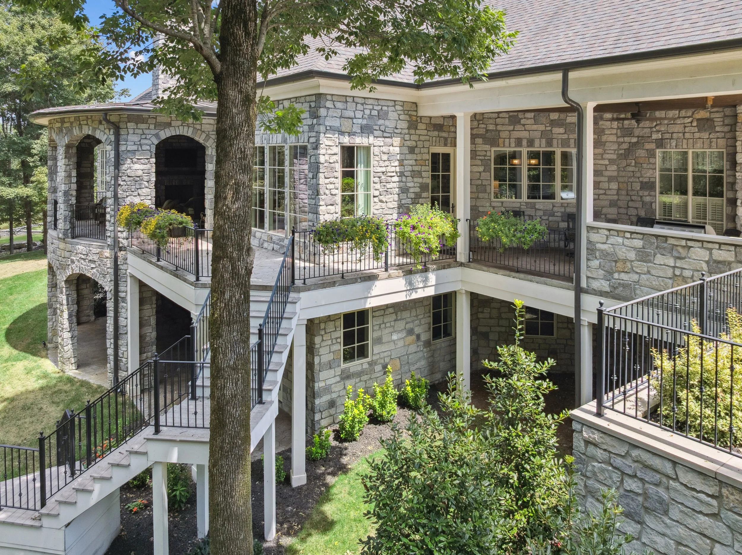 The exterior of a multi-story home with stone walls, large windows, a balcony with flower pots, and a staircase surrounded by greenery and trees.
