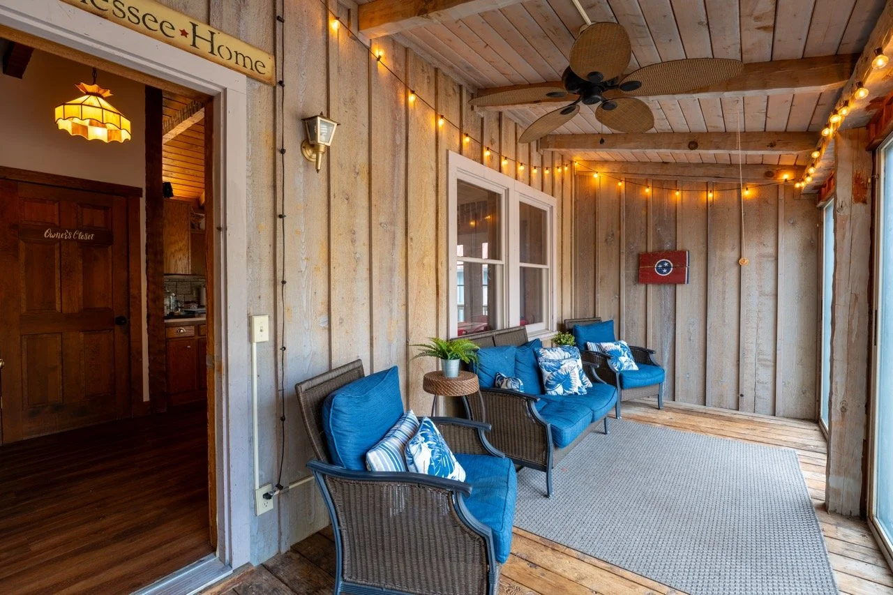 A cozy screened-in porch with wooden walls and ceiling, decorated with string lights, ceiling fan, and navy blue cushioned outdoor chairs with patterned pillows, a small side table with a plant, and a _Tennessee_ flag on the wall.