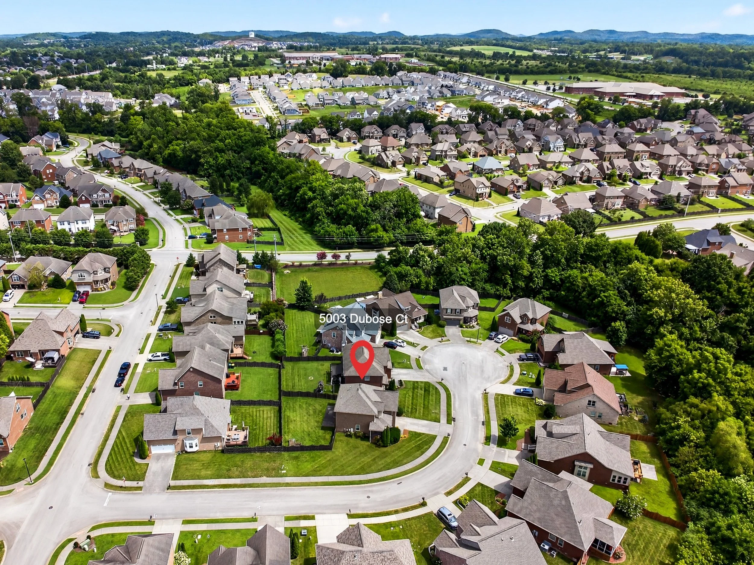 Aerial view of a suburban neighborhood with houses, green lawns, trees, and winding streets in a scenic area with hills and distant landscape.