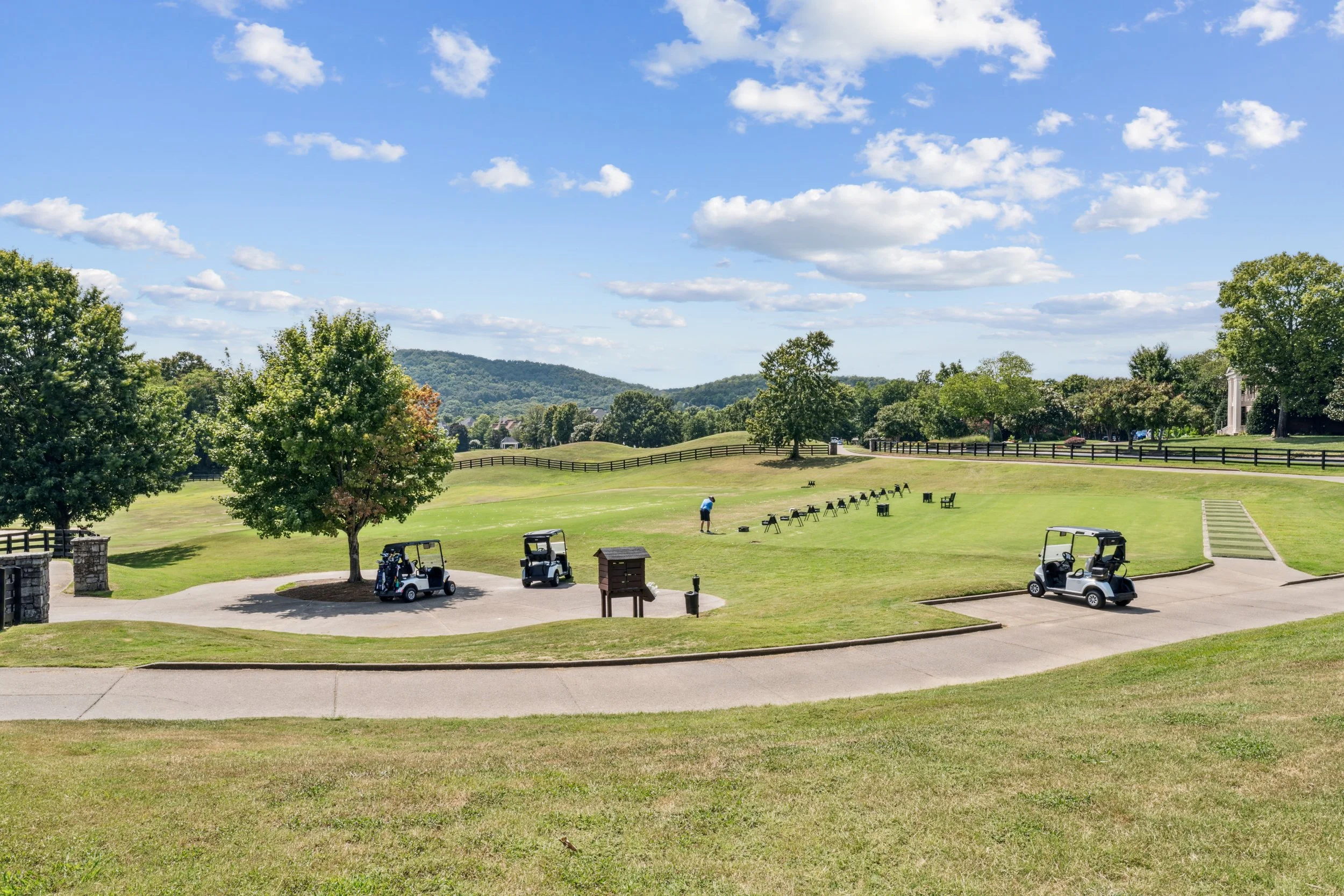 A golf course with several golf carts parked near the path, a person practicing golf swings, trees, benches, and a railing overlooking a hill, under a blue sky with scattered clouds.