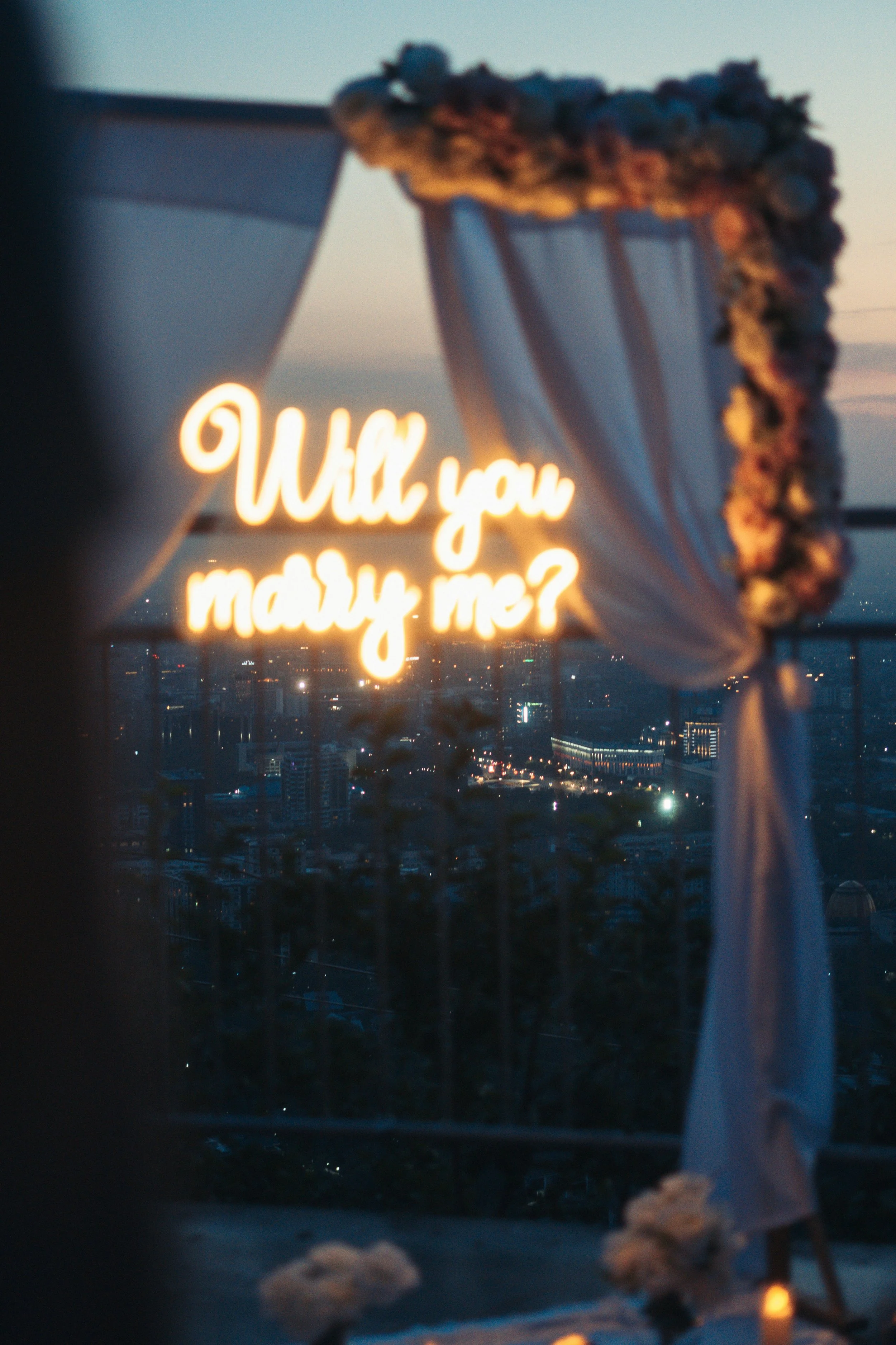 Luxury rooftop proposal setup with fabric drapes, warm candlelight and LED ‘Will You Marry Me?’ sign, styled by Whispers of Yes in Essex.