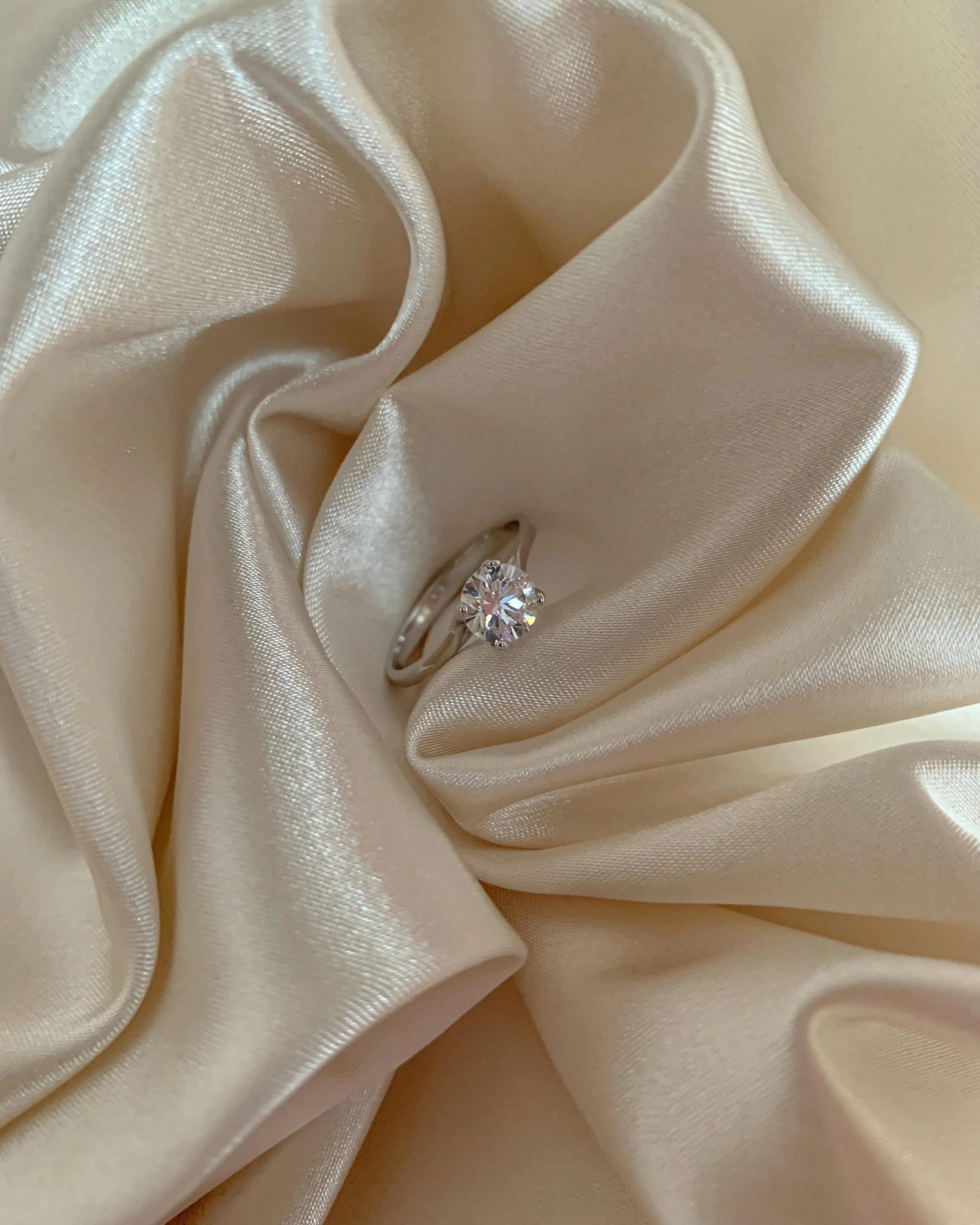 Diamond engagement ring styled on ivory satin fabric, timeless proposal detail captured by Whispers of Yes