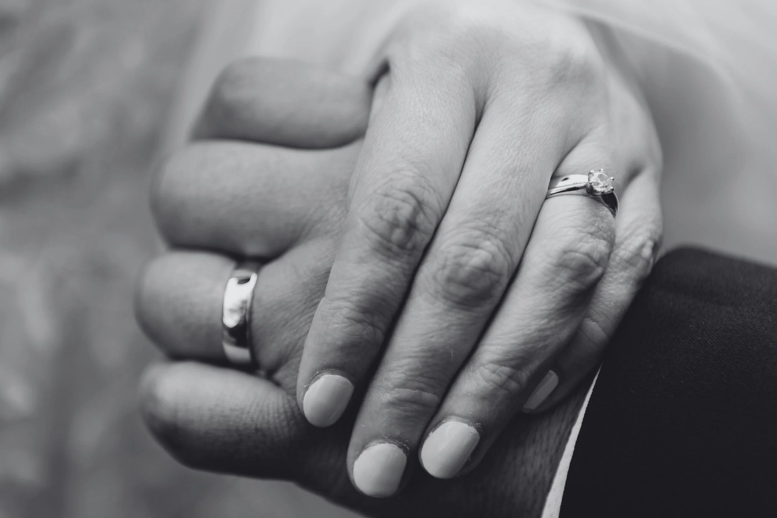 Black-and-white close-up of clasped hands after a Whispers of Yes proposal, wedding rings and emotion captured in soft detail.