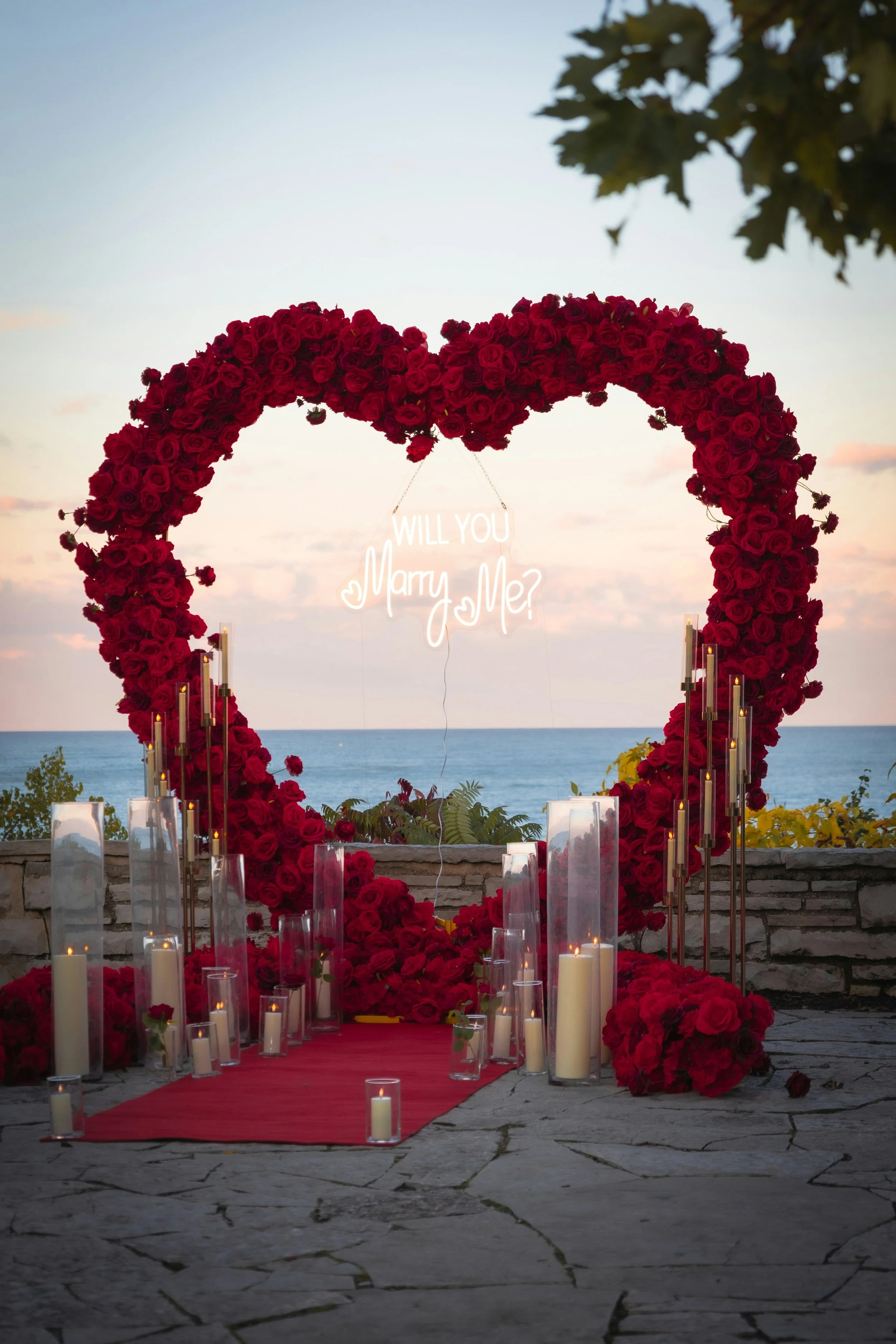 Luxury heart-shaped red rose arch with ‘Will You Marry Me?’ neon sign, romantic outdoor proposal setup with candlelight and premium décor