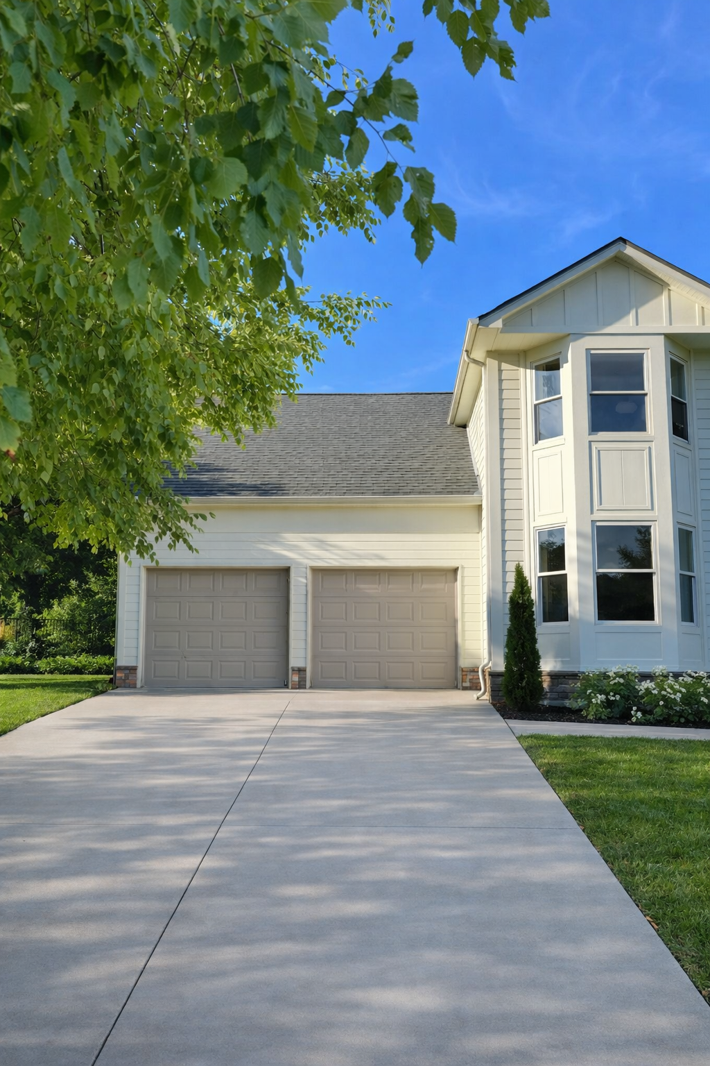 A house with a concrete driveway, white exterior, bay windows, and a two-car garage in a suburban neighborhood, with a green tree and a clear blue sky.