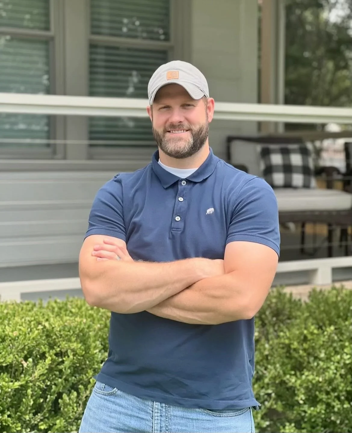 A man with a beard wearing a light gray baseball cap and a navy blue polo shirt, standing confidently with arms crossed in front of a house with a porch and greenery.