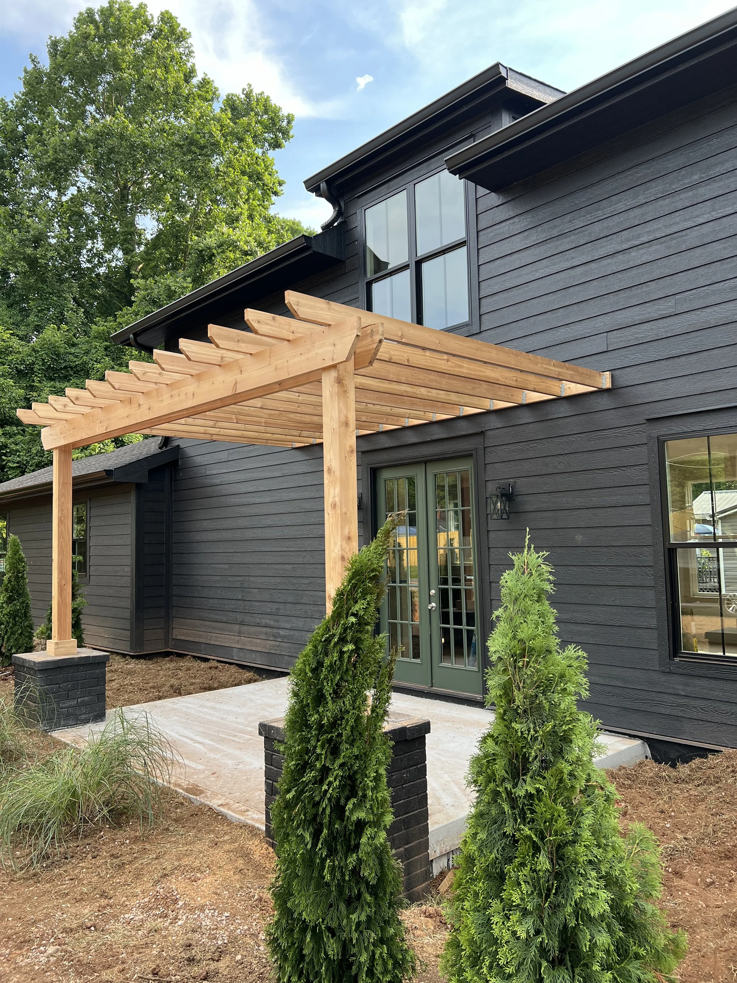A black wooden house with a modern design features large windows and glass doors. In front, there is a small patio area with a newly constructed wooden pergola. Several green shrubs planted in black brick planters line the patio, and the yard has bare soil, indicating ongoing landscaping. Tall trees are visible in the background, and the sky is partly cloudy with sunlight.