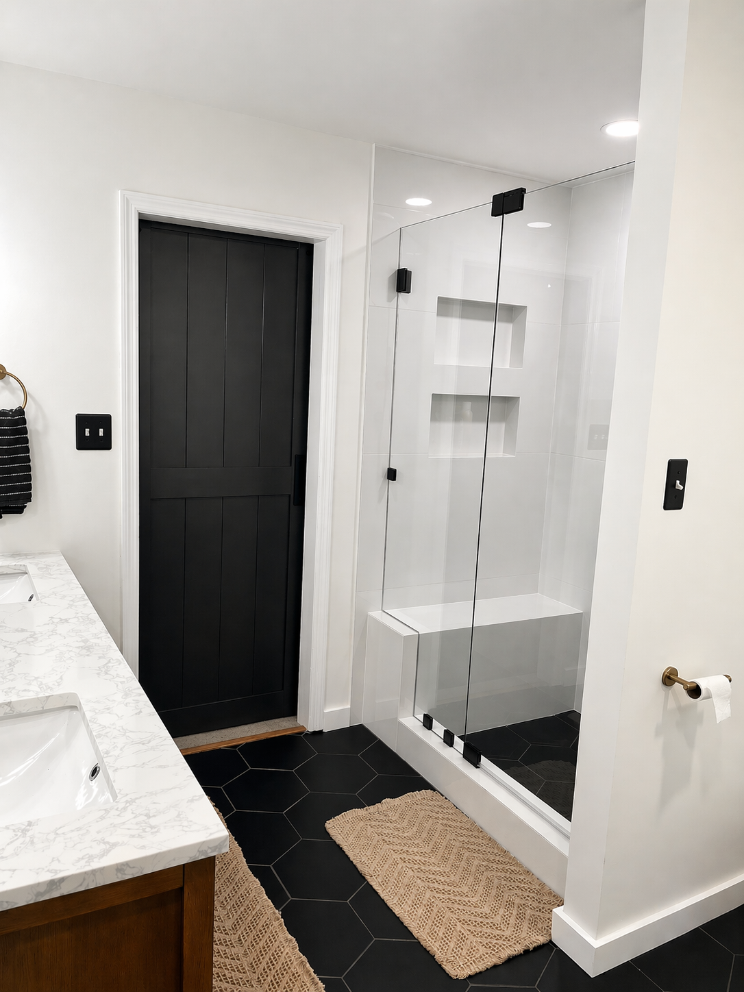 Modern bathroom with a black sliding door, glass shower enclosure with built-in shelves, black hexagon floor tiles, beige bath mats, part of a marble countertop with dual sinks, and white walls.