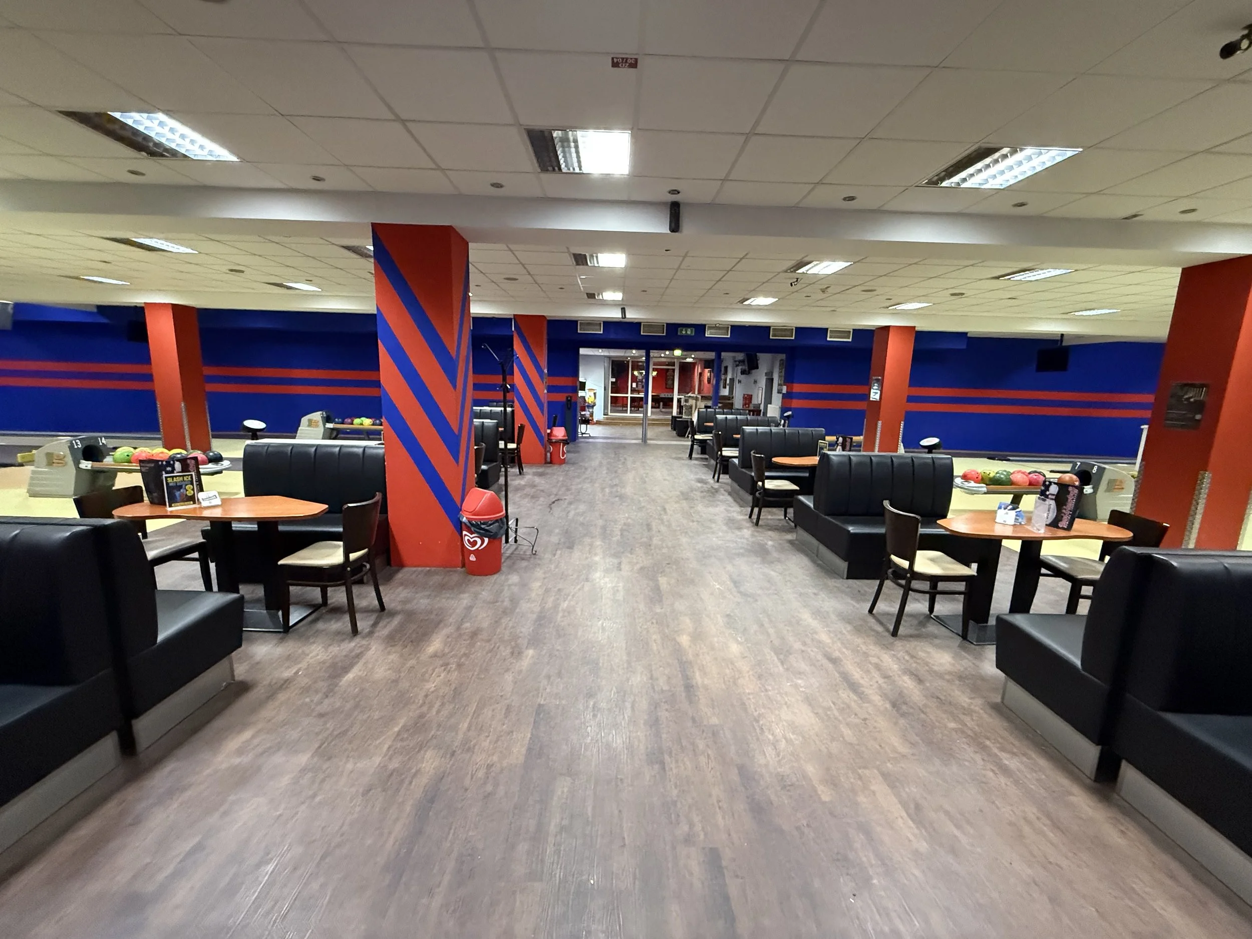 Leeres Bowlingcenter mit schwarz gepolsterten Sitzbänken, Tischen und Bowlingbahnen, bunte Kugeln und Gänge im Hintergrund.