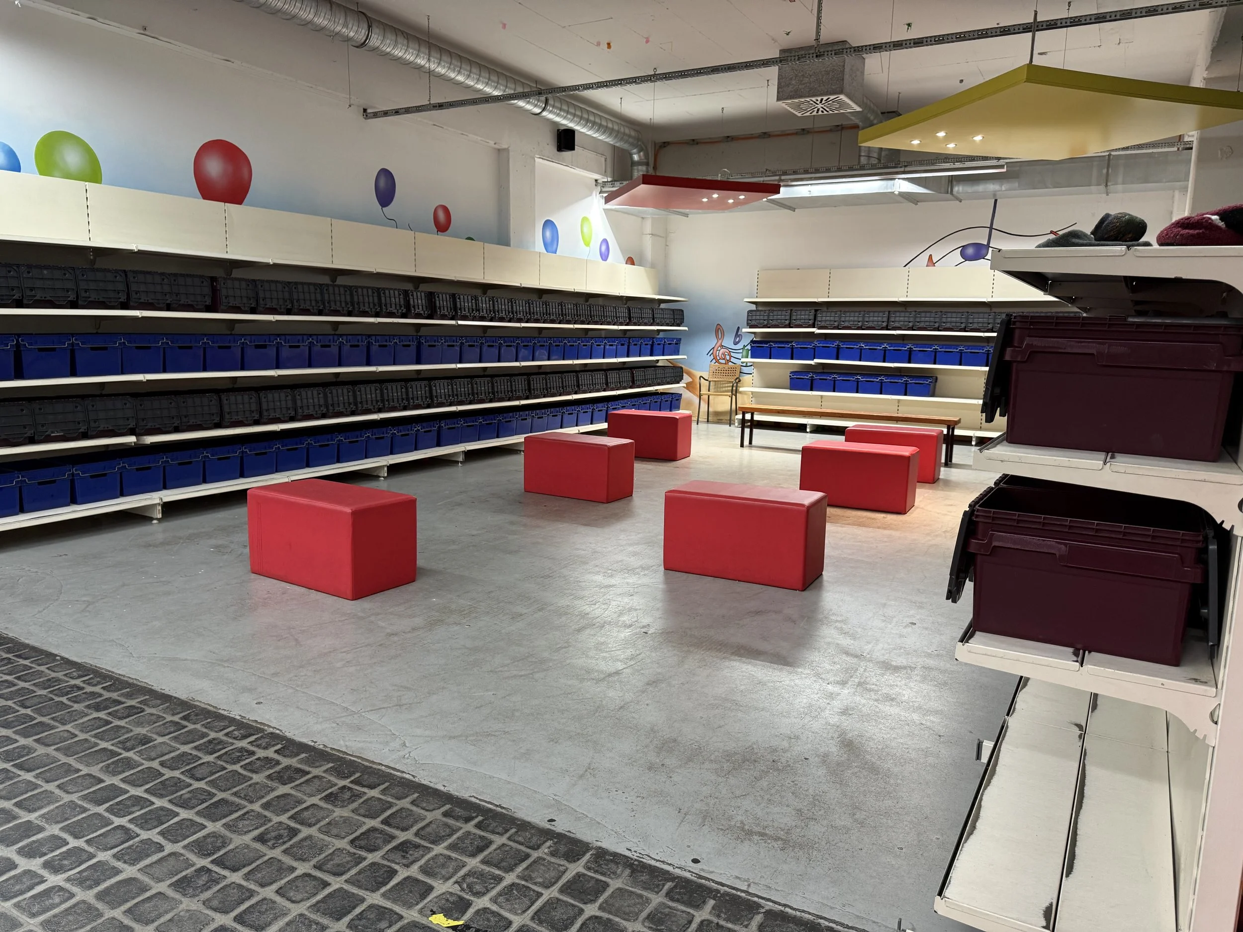 Leerer Ladenraum mit roten Sitzwürfeln, leeren Regalen mit blauen und schwarzen Behältern, bunten Ballon-Dekorationen an der Wand, und einer Sitzbank. Zur Dekoration eine bunte, musikalische Note auf der Wand.
