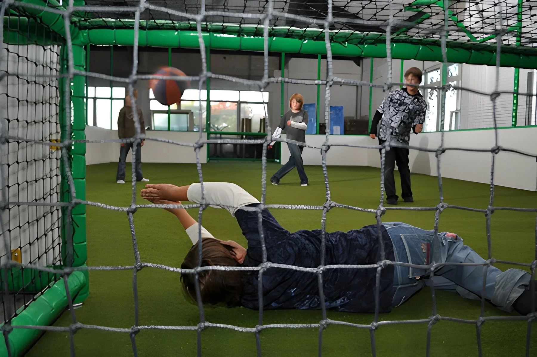 Junge liegt auf dem Boden hinter einem Netz und versucht, einen Basketball zu fangen oder zu blockieren, während andere Personen im Innenumfeld spielen.