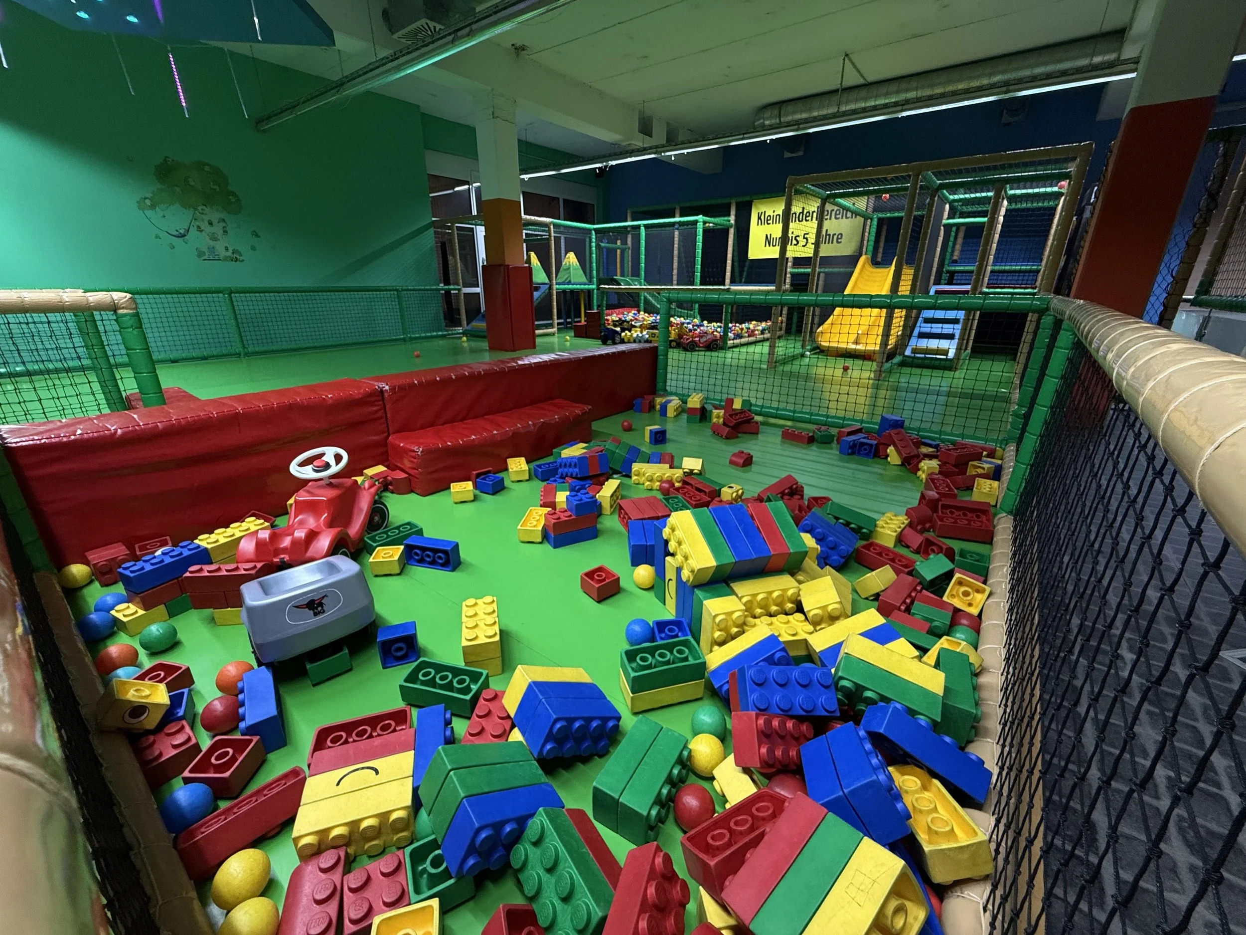 Ein Indoor-Spielbereich mit einer Rutsche, umgeben von bunten LEGO-Steinen, Bällen und Klettergerüsten. Es ist ein Kinderspielplatz mit weichen Matten und Spielstrukturen.