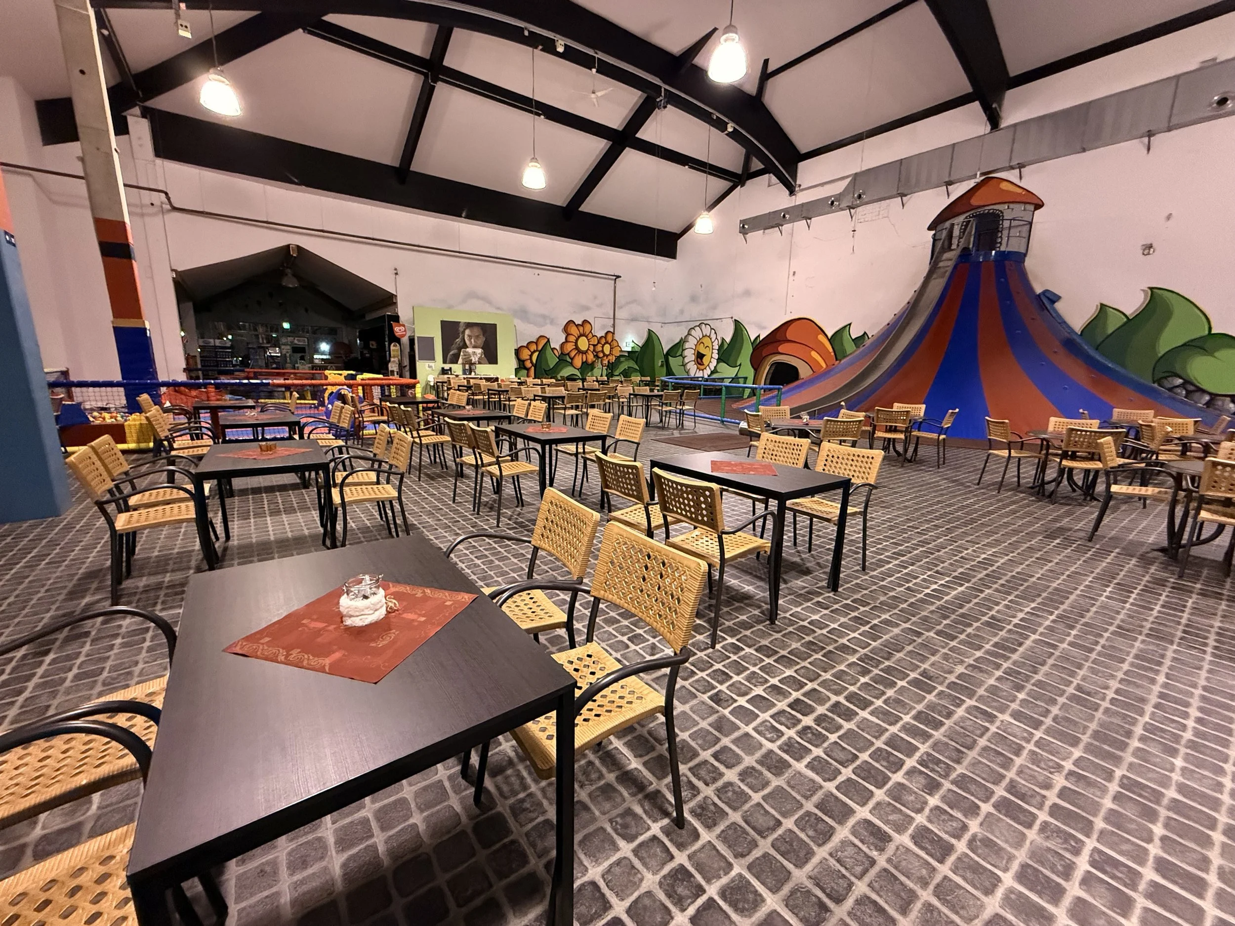Ein leeres Indoor-Spielplatz mit Tischen und Stühlen, bunten Wandmalereien und einer großen Rutsche im Hintergrund.