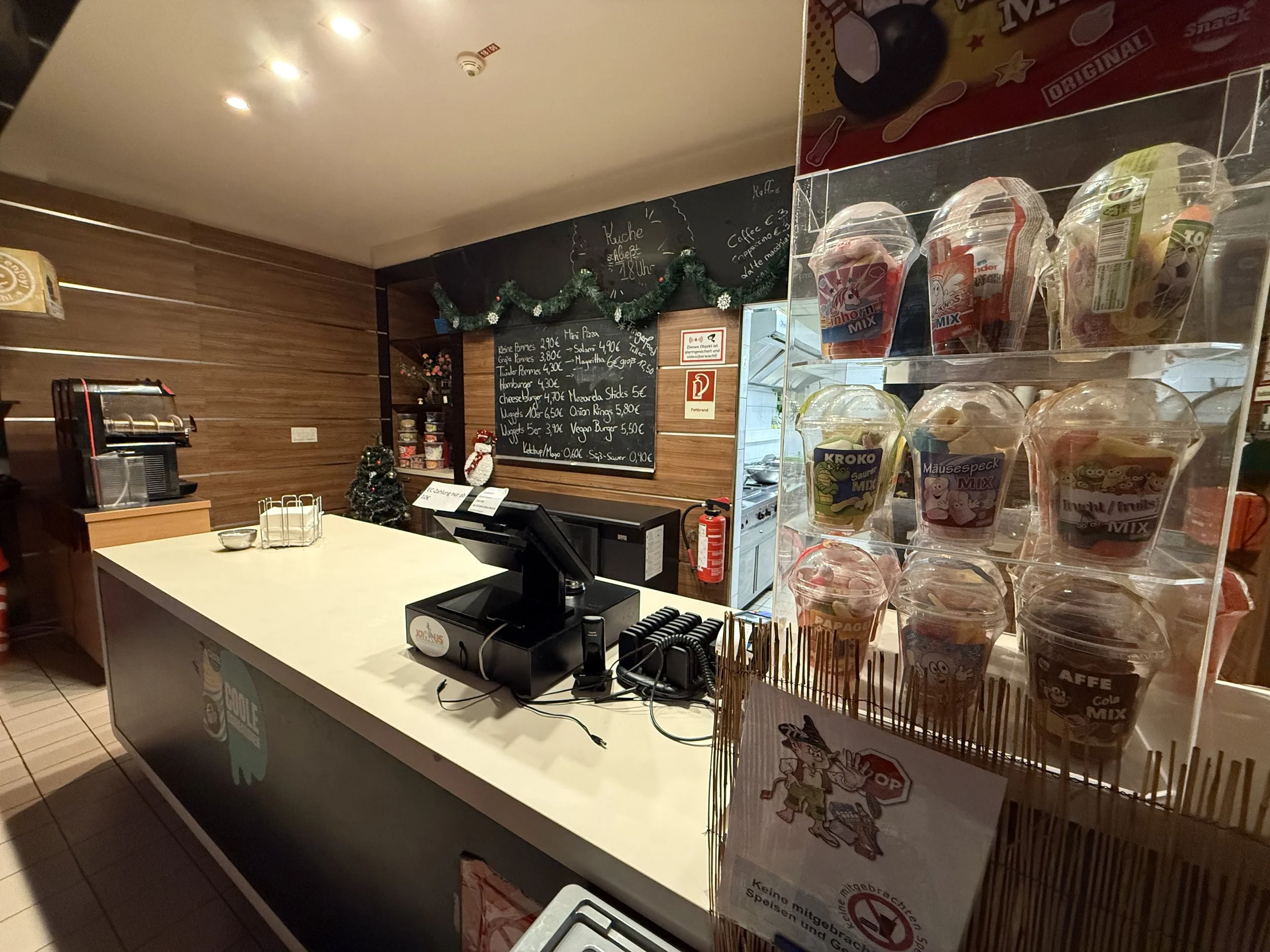 Innenaufnahme eines Imbiss- oder Kiosk-Thekenbereichs mit weißen Theken, einer digitalen Kasse, einem schwarzen Kasten mit Zutaten und einem Regal mit bunten Eisbechern in transparenten Bechern, auf dem rechten Bildrand. Im Hintergrund eine Wand mit einer Tafel, auf der Speisen und Preise in Schwarz geschrieben sind, und eine kleine Weihnachtsdekoration, darunter eine Mini-Weihnachtsbaum. Weihnachts- und Dekorationsartikel sind auf der Theke zu sehen.