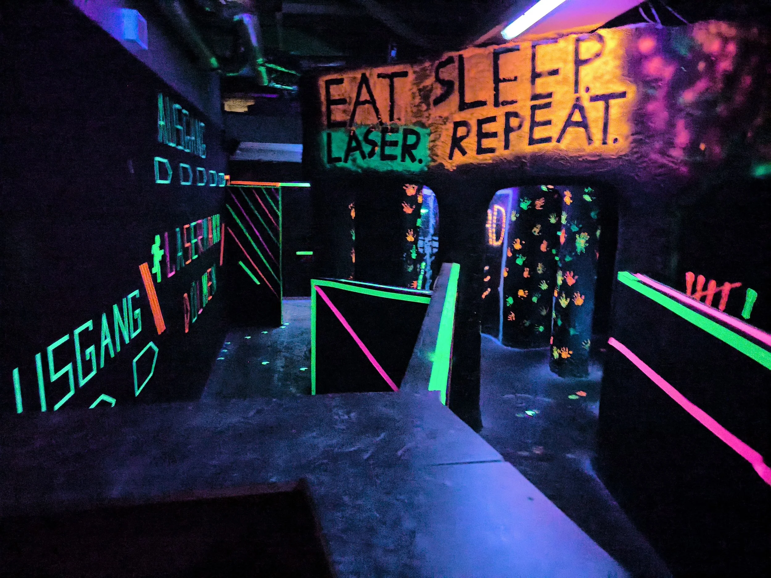 Dunkler Raum mit Neonlichtern, einer Wand mit der Aufschrift 'Essen Schlaf Laser Repeat' und einem dekorierten Vorhang mit leuchtenden Pflanzmustern.