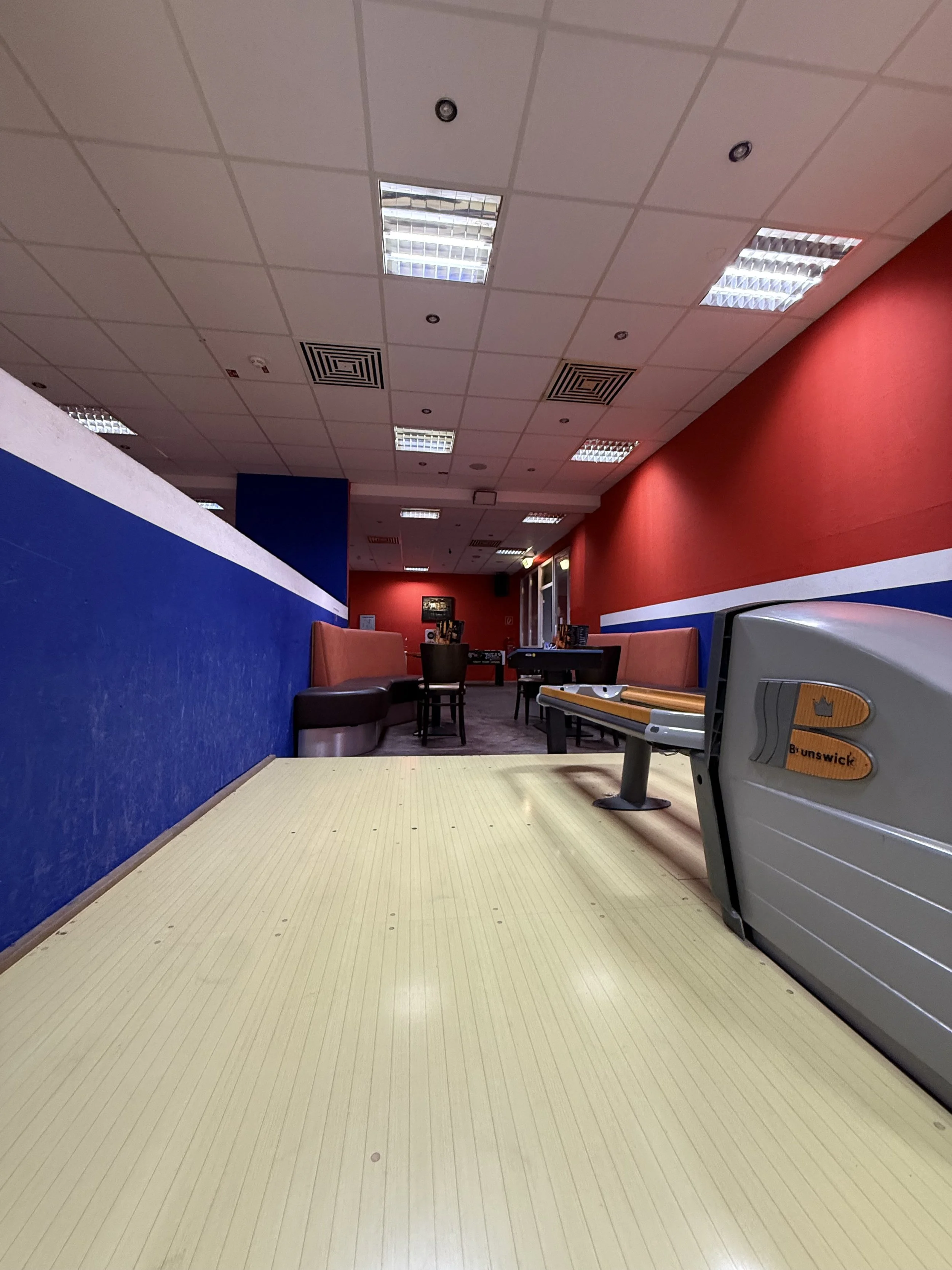 Innenansicht eines Bowlingbahns mit Sicht auf den Sitzbereich mit Tischen und Stühlen, rote und blaue Wände, Deckenleuchten