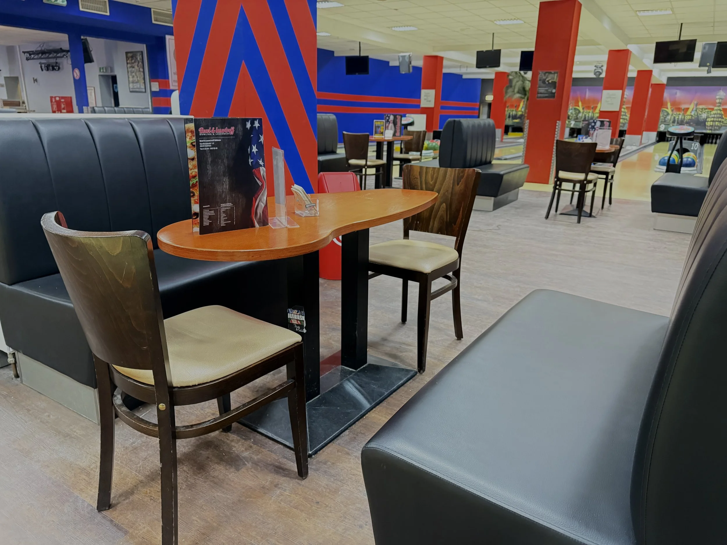 Leeres Bowling-Areal mit Sitzgelegenheiten und einem Tisch im Vordergrund, bunte Wanddekorationen, mehrere Bowlingbahnen im Hintergrund.