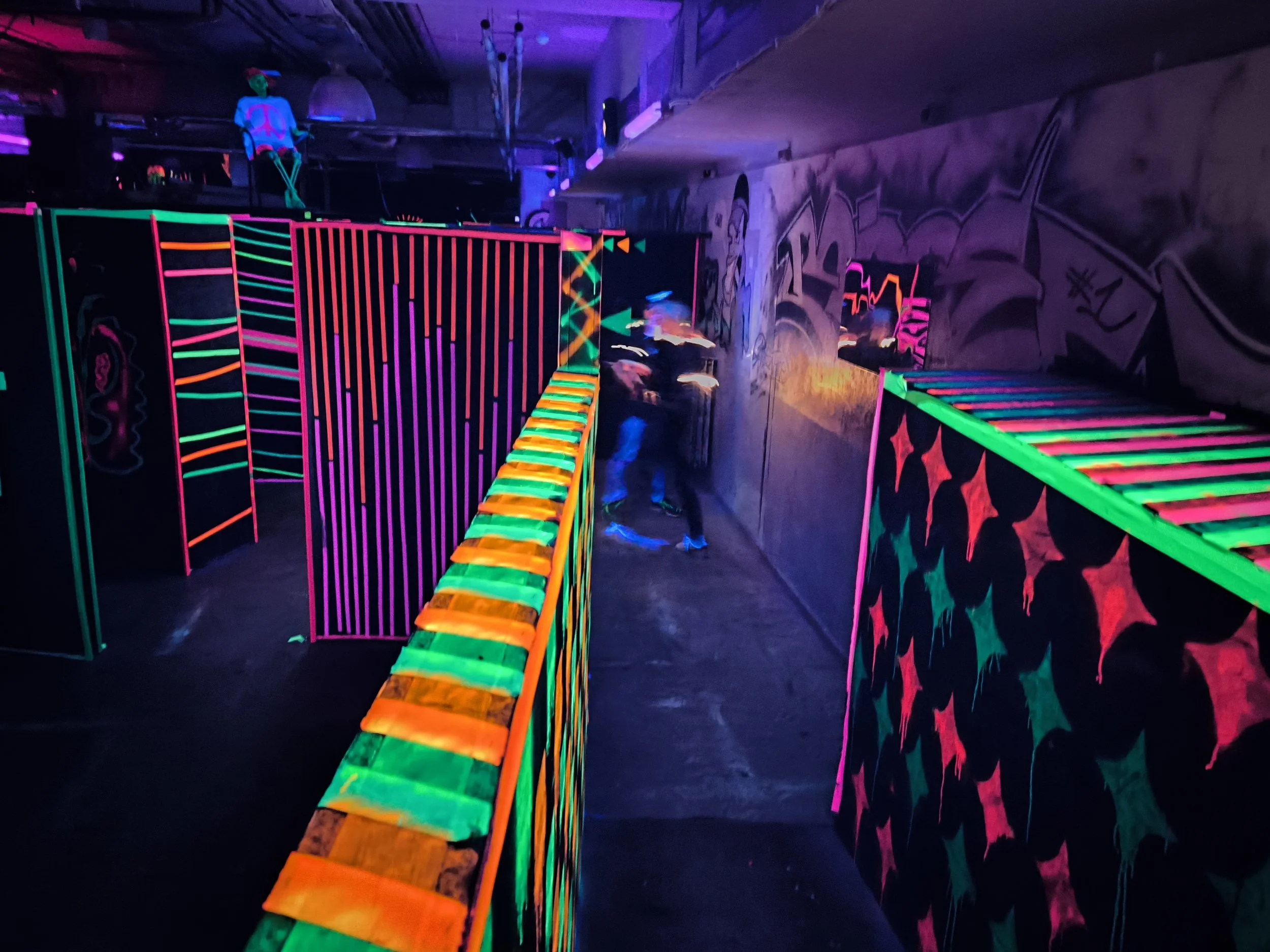 Eine Laser-Tag-Arena mit neonfarbenen Stangen und Wänden, beleuchtet mit Schwarzlicht, inklusive Person in dunkler Kleidung und einem Neon-Kostüm.
