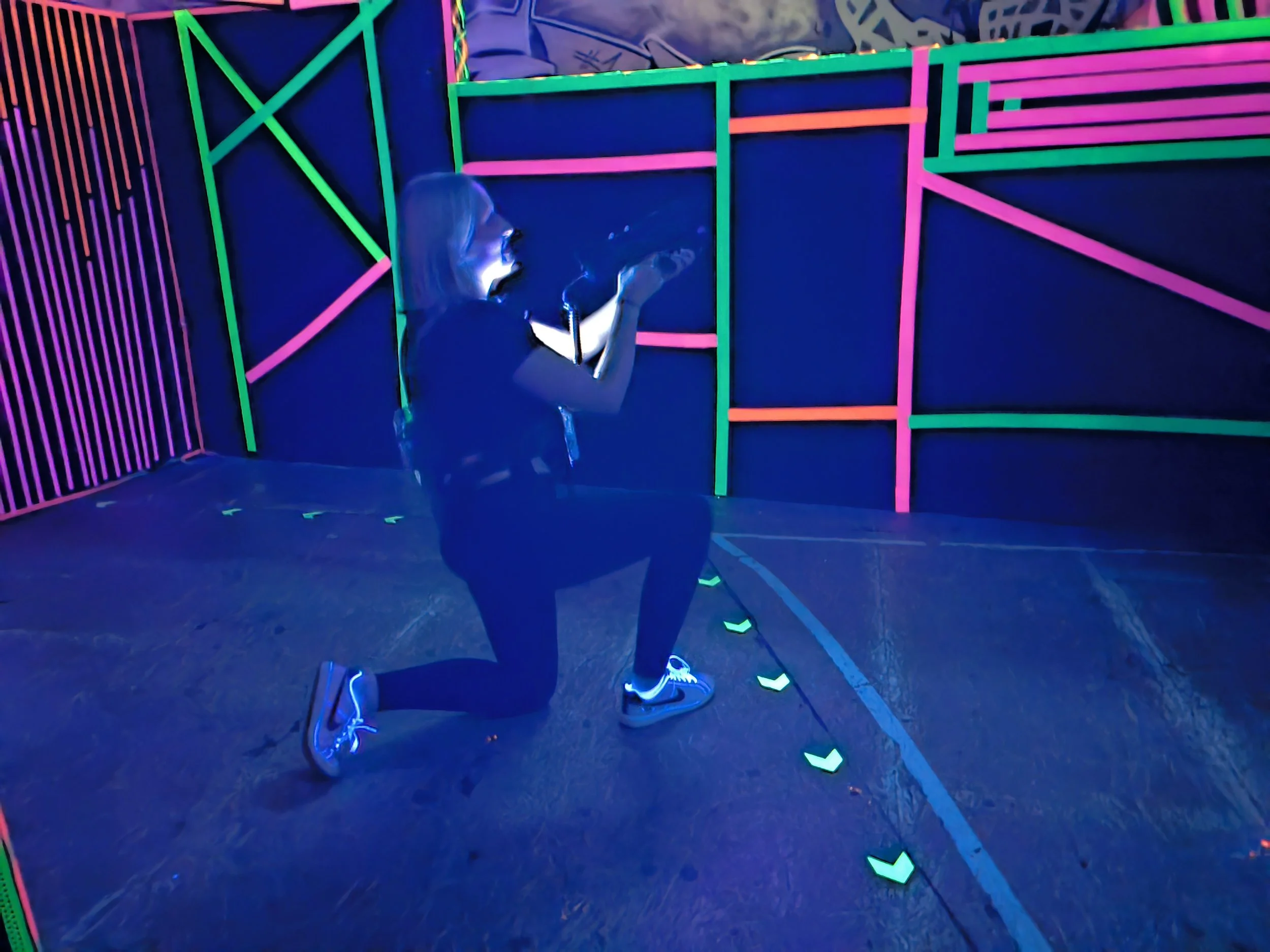 Eine Person im Dunkeln, die Neonlichter trägt, kniet auf einem Boden, umgeben von leuchtenden Linien und Rahmen in einem Neon-Labyrinth, das für ein Virtual-Reality-Spiel oder eine elektronische Kunstinstallation gestaltet ist.