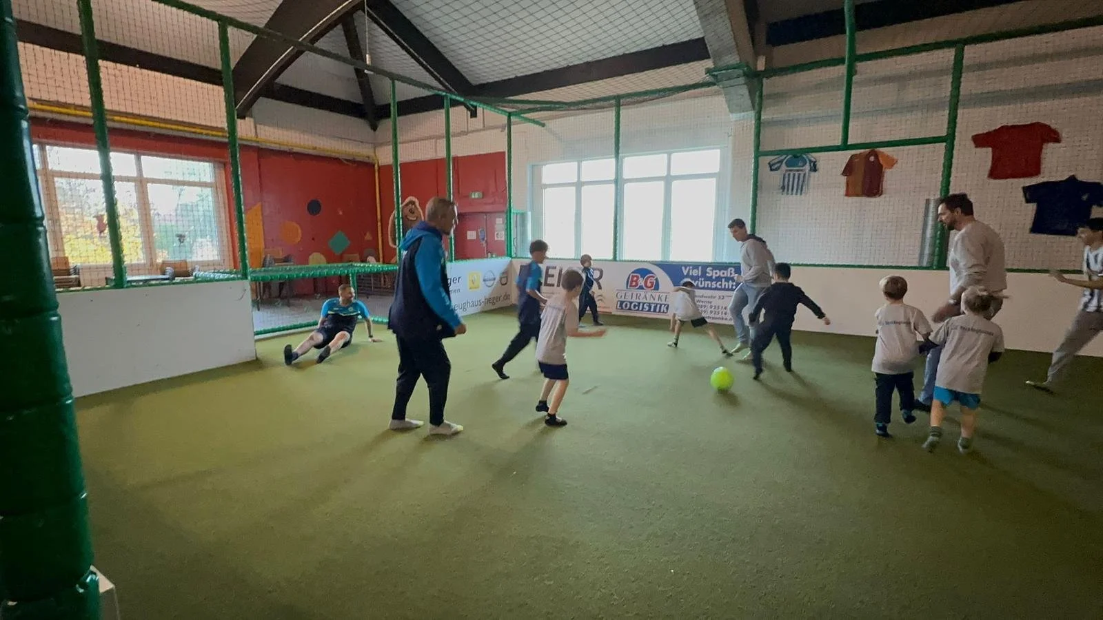 Junge spielen Fußball in einer Indoor-Halle, begleitet von Erwachsenen.