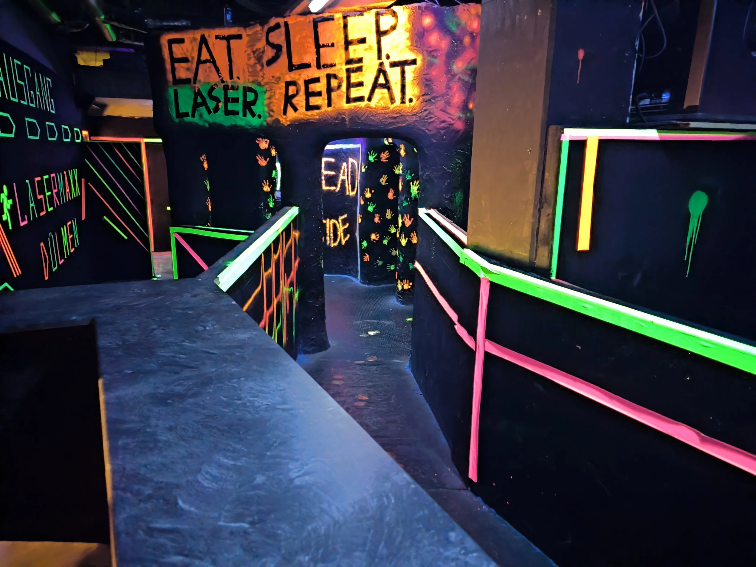 Neonbeleuchtete Laser-Tag-Indoor-Spielhalle mit bunten Wänden, die mit fluoreszierender Schrift und Handabdrücken dekoriert sind. Das Wanddesign erinnert an ein Street-Art- oder Graffiti-Stil. Neonleuchtende Schilder mit Texten wie 'Eat Sleep Laser R