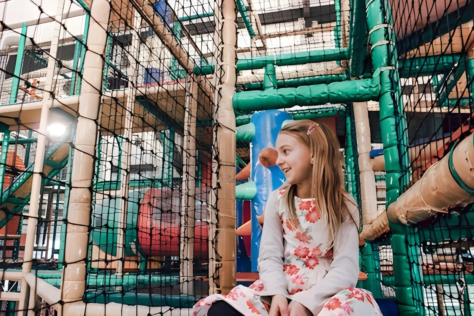 Ein junges Mädchen sitzt in einem Kinderspielplatz mit einem Klettergerüst aus Holz und Plastik, umgeben von Netzen und Rohren.