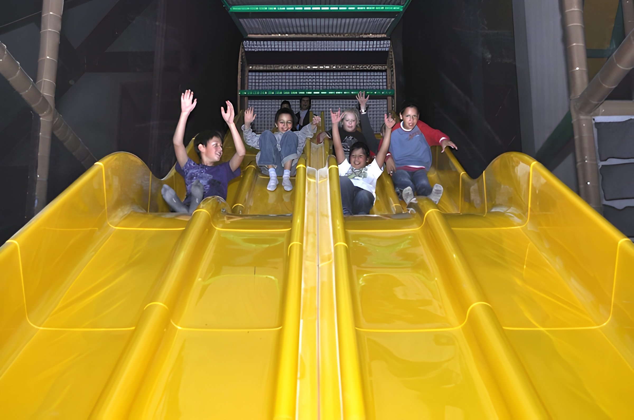 Fünf Kinder rutschen auf einer großen, gelben Rutsche im Indoor-Spielplatz und haben Spaß.