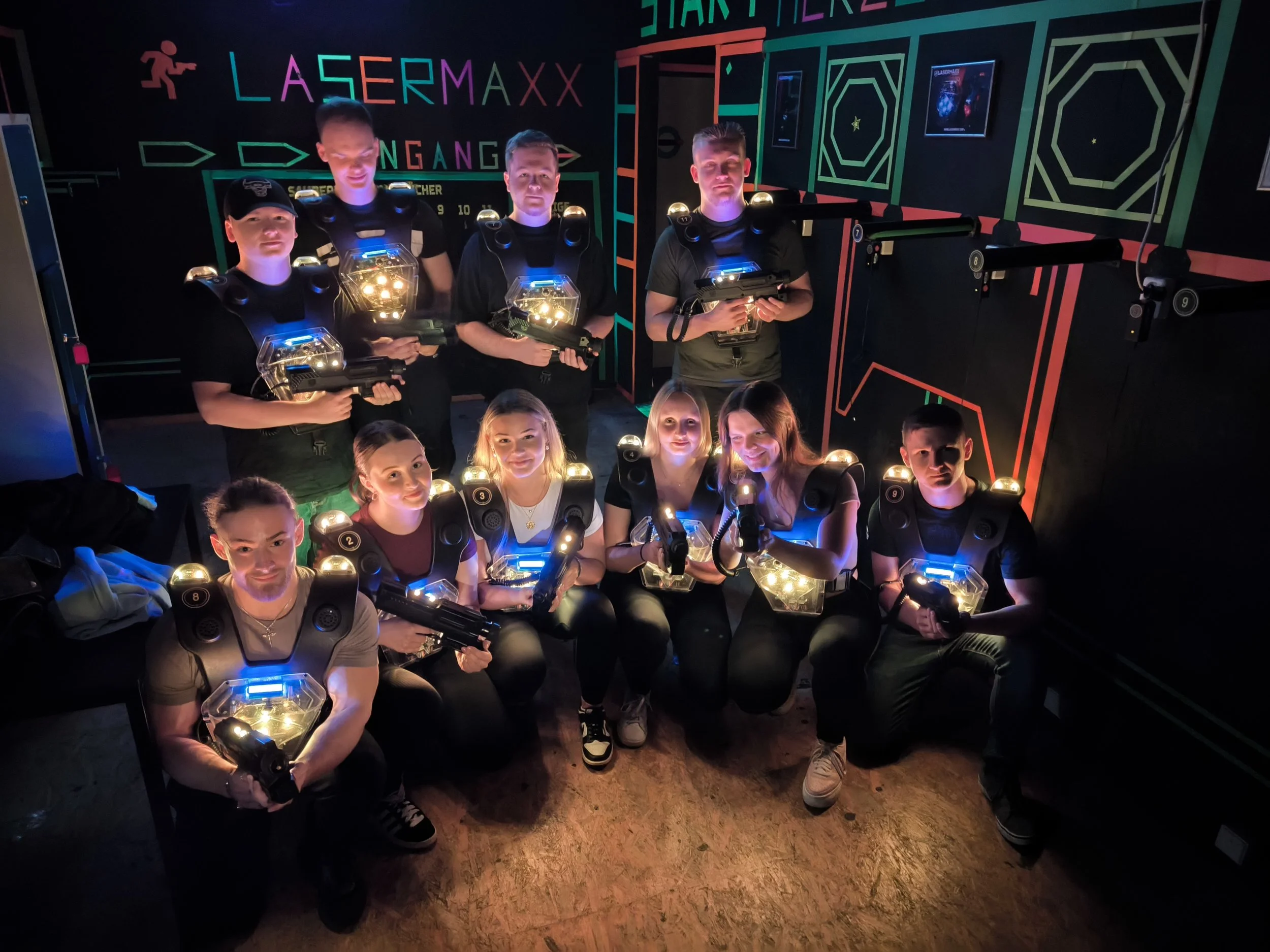 Gruppe von acht Menschen in einer Laser-Tag-Arena, alle tragen Laser-Helme und halten Lasergewehre, die im dunklen Raum mit bunten Neonlichtern leuchten.