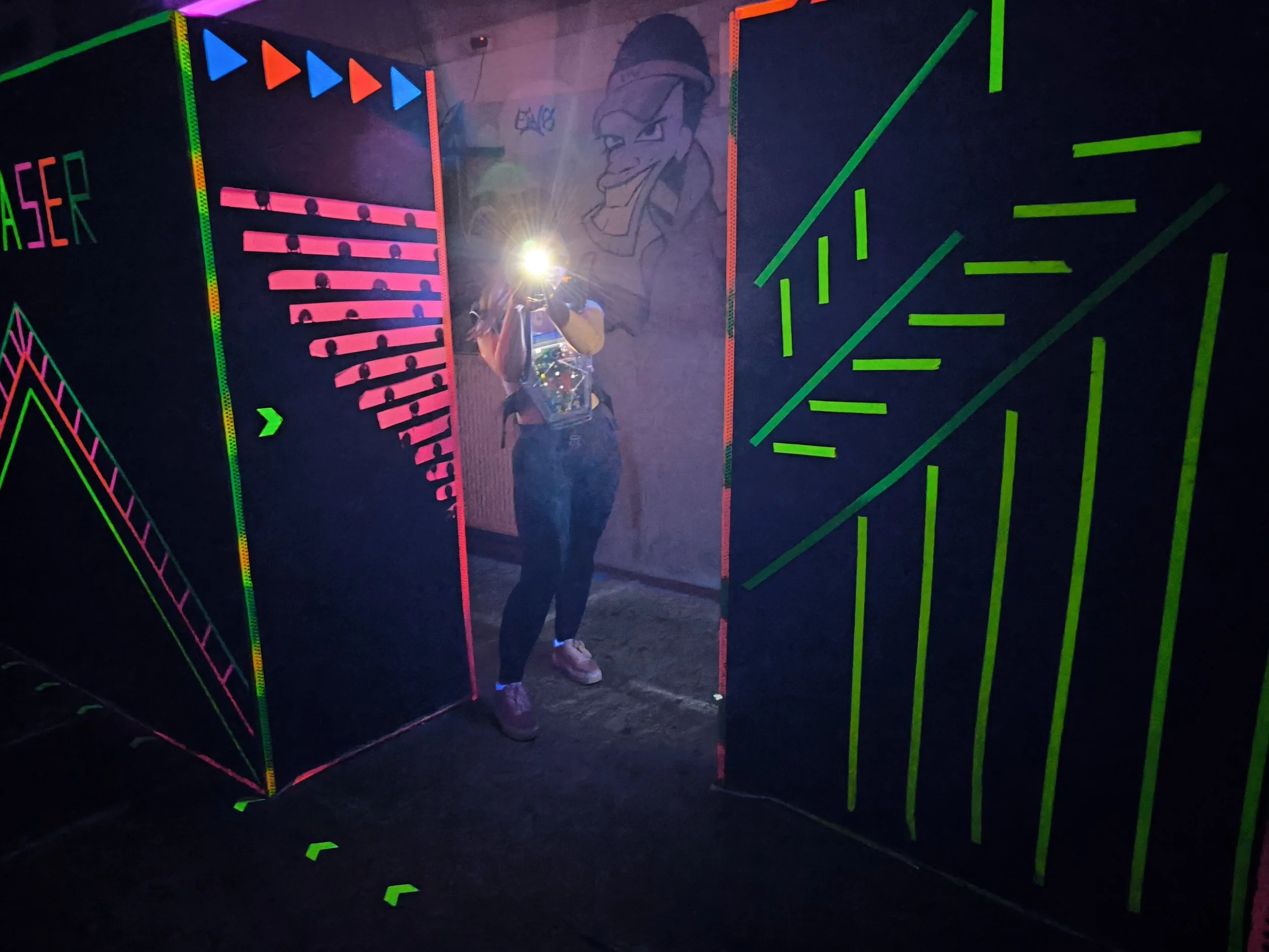 Person mit Rucksack und Kamera in einer bunten Schwarzlicht-ähnlichen Umgebung mit geometrischen Mustern und einer Wandzeichnung eines lachenden Mannes im Hintergrund.